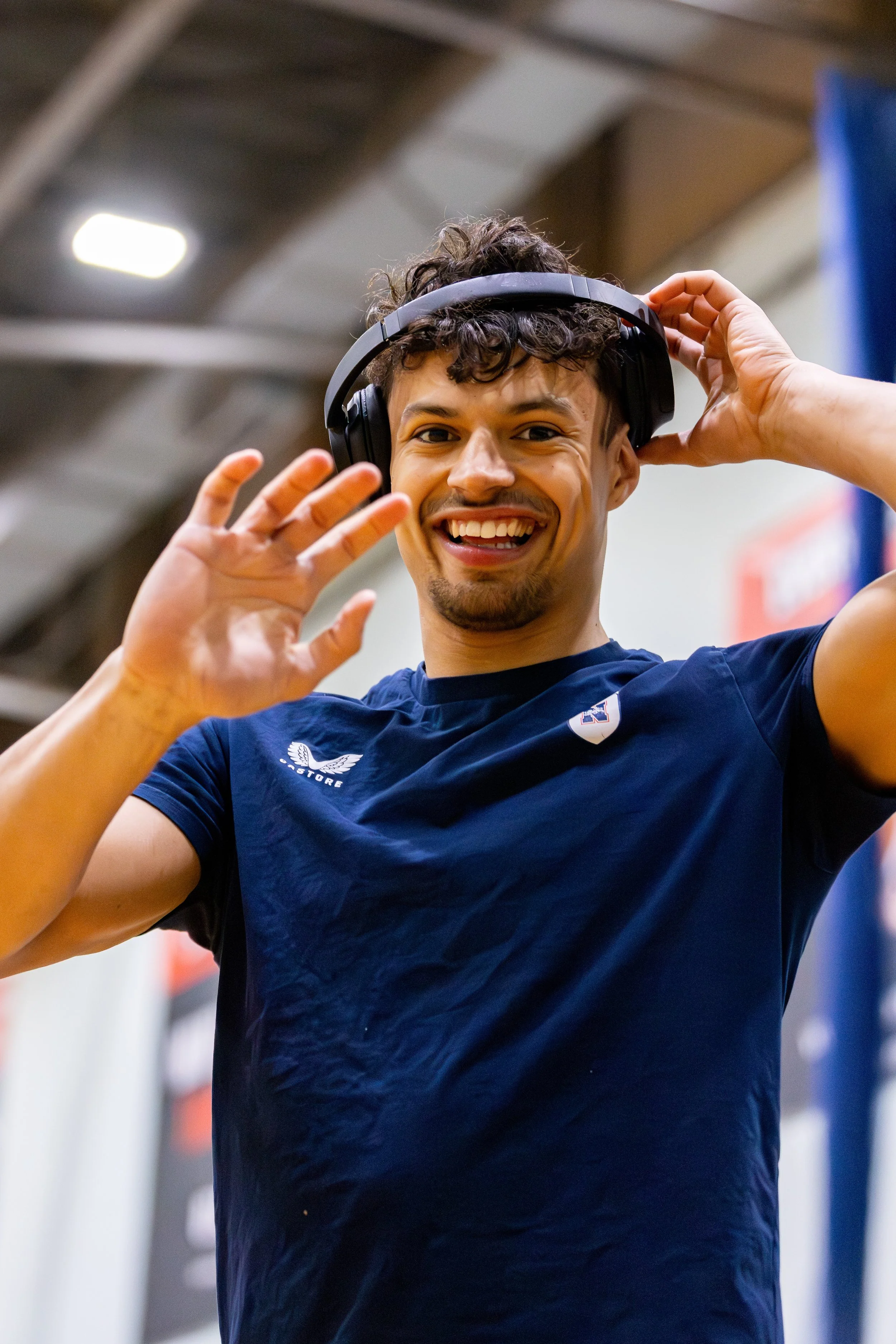 A young man with curly hair smiling and waving while putting on headphones in an indoor sports facility.