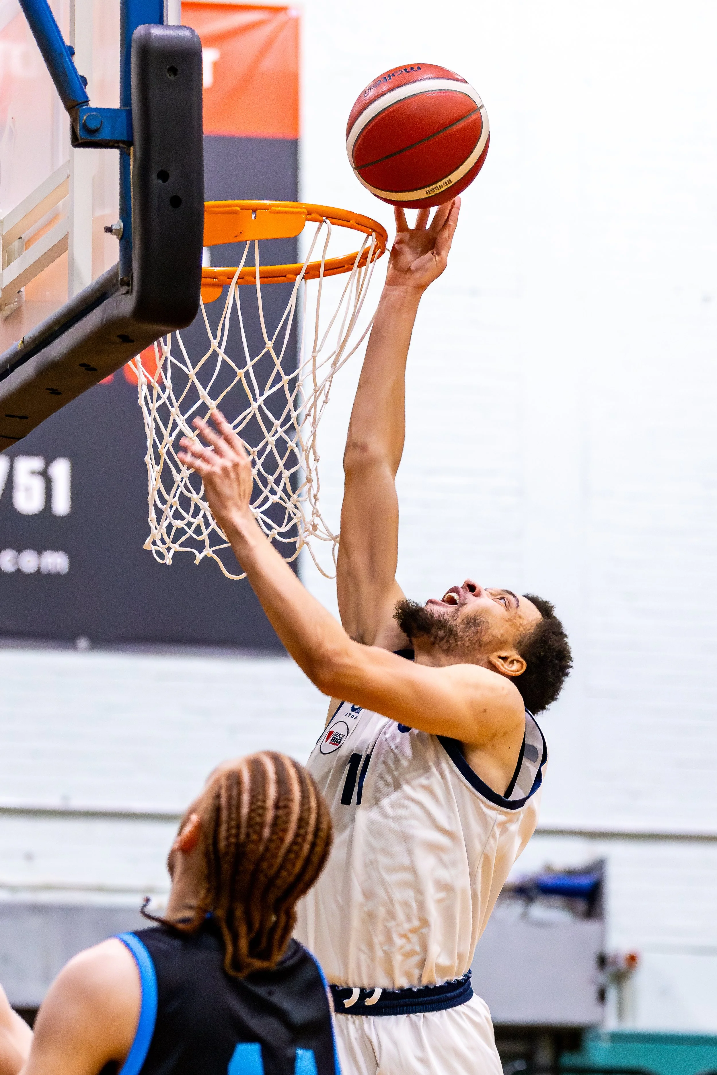 A basketball player in a white jersey is jumping to his left to make a shot at the basket, with another player in a black jersey watching nearby.