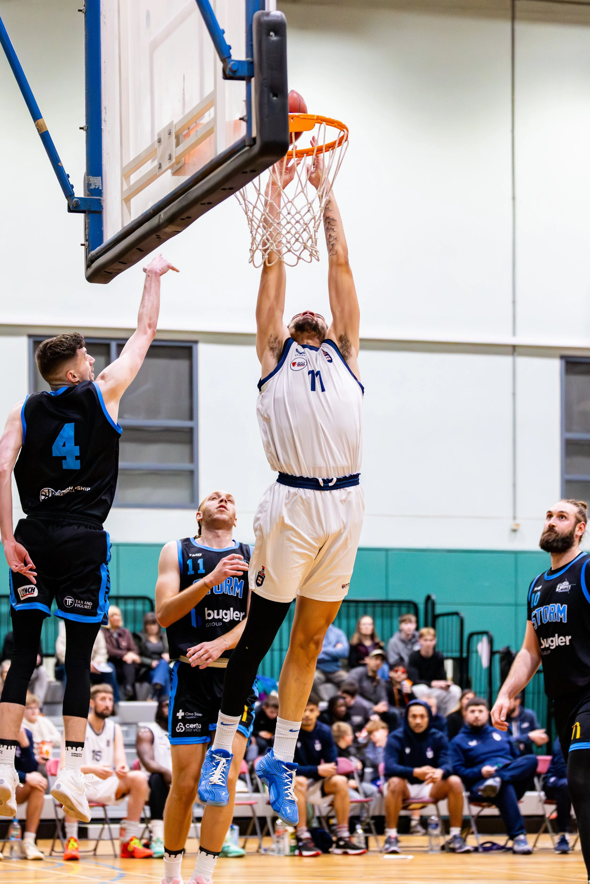 A basketball player in a white jersey jumping to score a basket while an opposing player in a black jersey attempts to block. Other players and spectators are visible in the background.