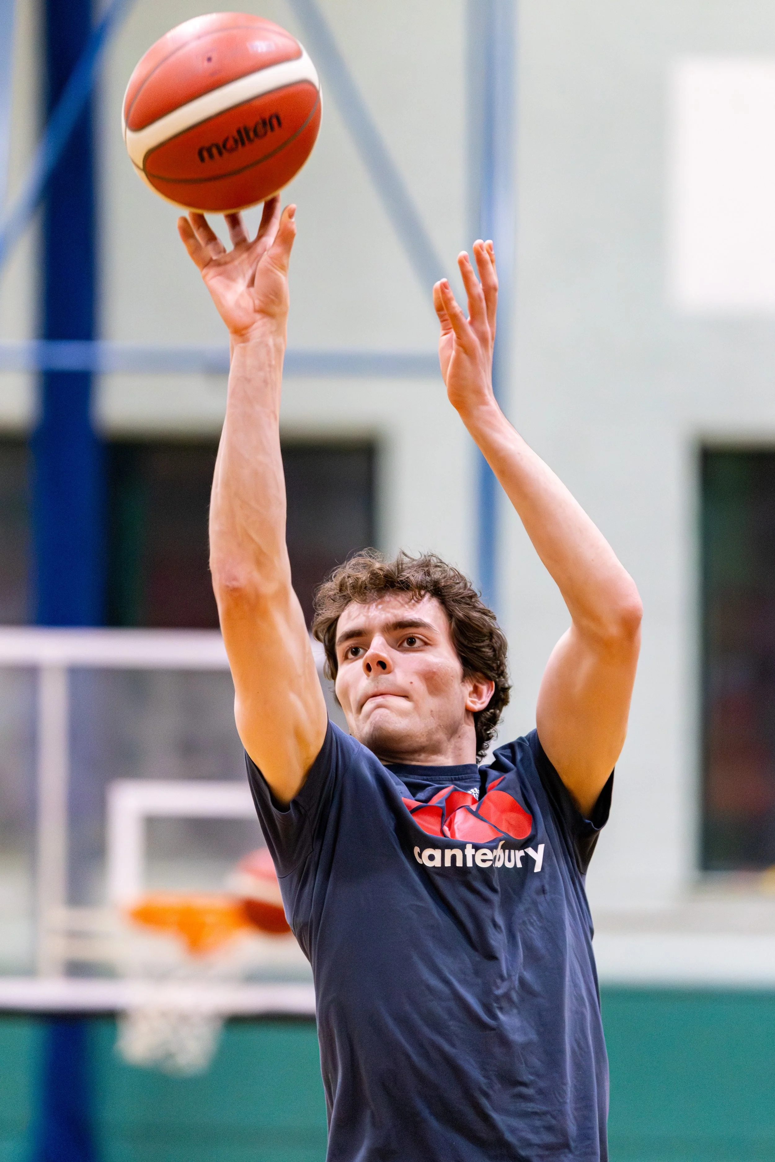 A male basketball player in a blue shirt with the word 'Canterbury' is jumping to shoot a basketball during a game, with a focused expression on his face.