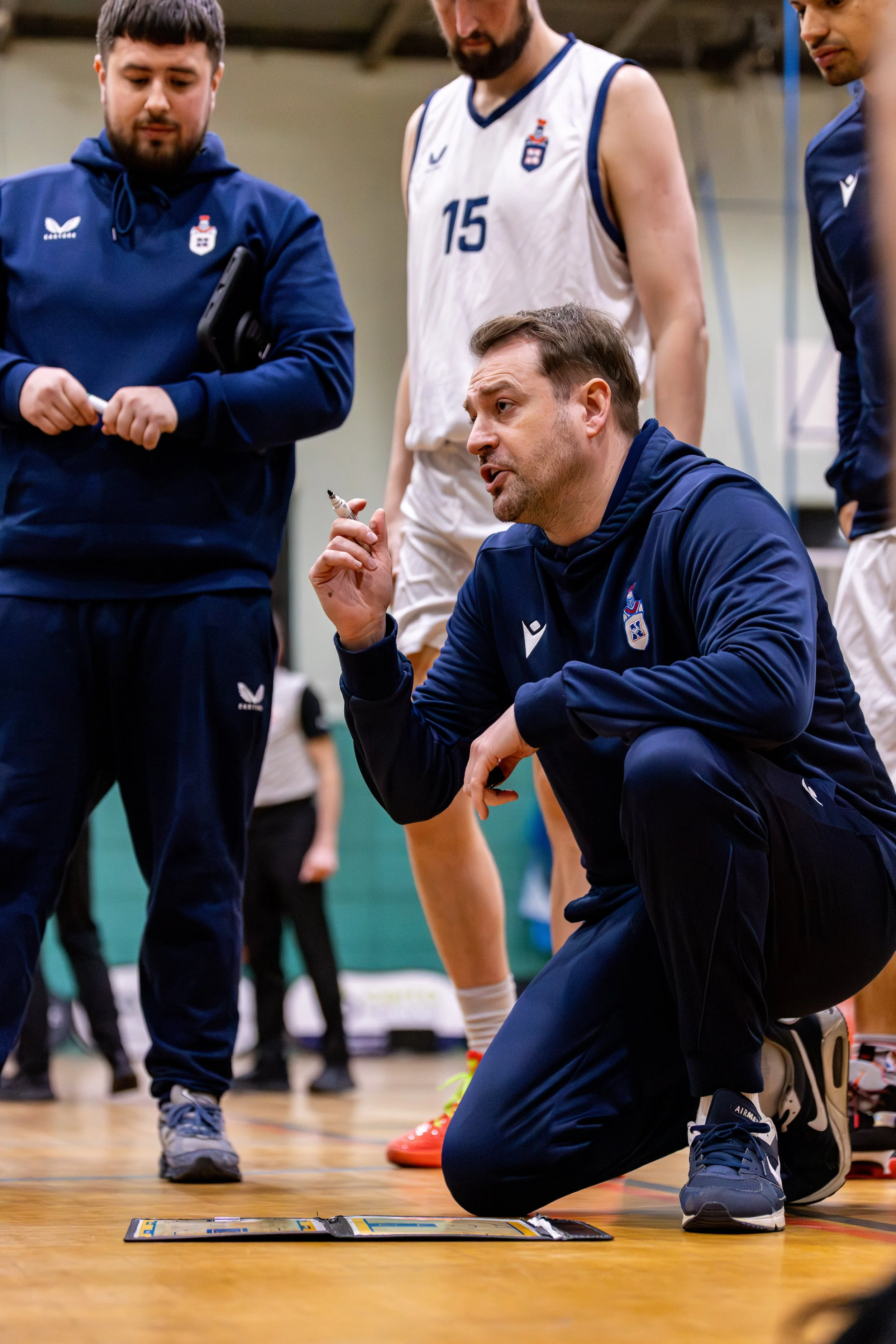 A basketball coach kneeling on the court floor during a game, giving instructions to his team, with players standing around him, wearing team uniforms.