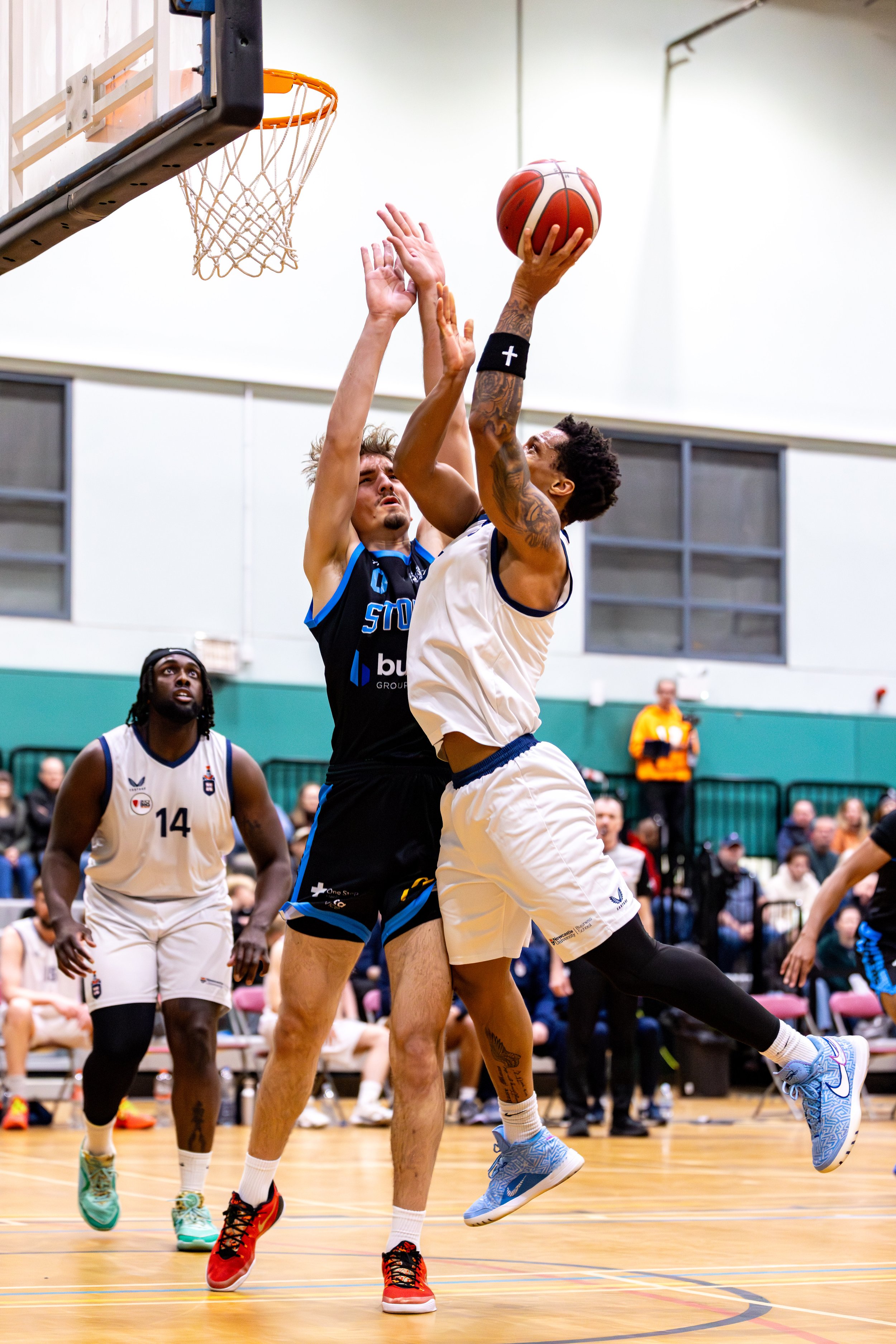 Two basketball players compete near the basket, one in a white jersey jumping with the ball and the other in a black jersey blocking. A third player in a white jersey watches nearby.