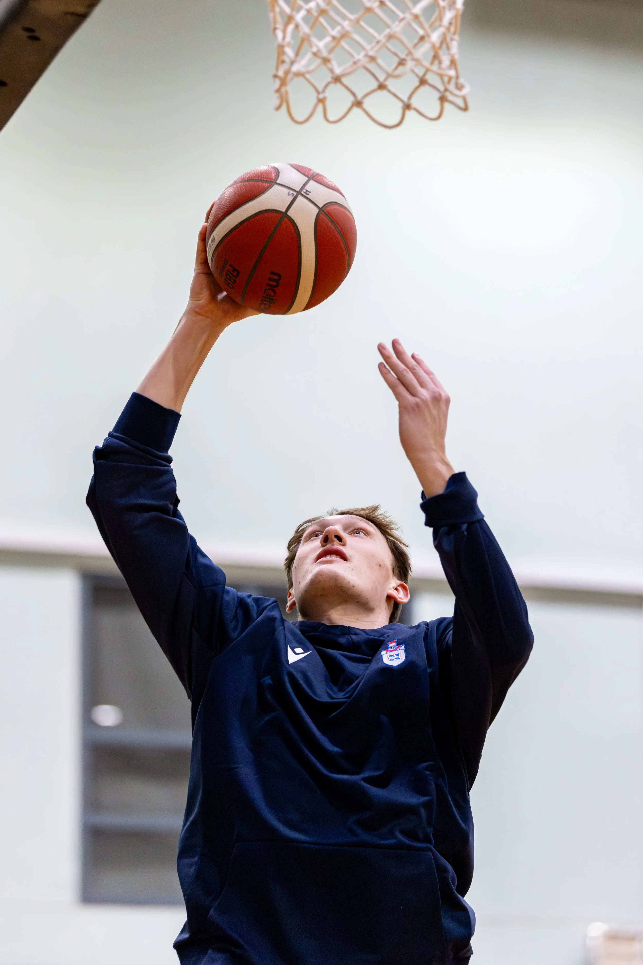 Young male basketball player wearing a navy blue jersey, about to shoot a basketball toward the hoop inside a gymnasium.