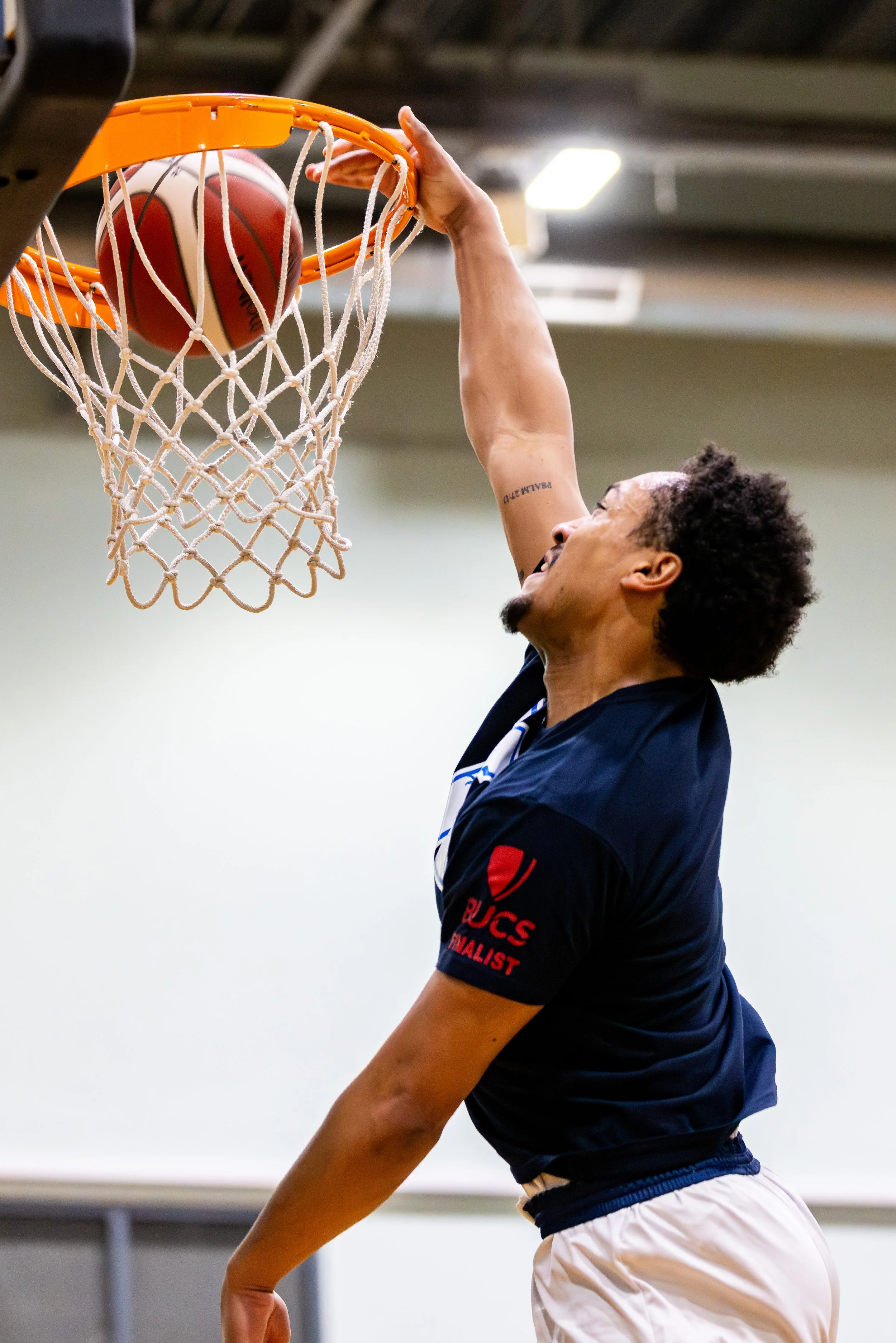 A basketball player is dunking a basketball into the hoop with one hand, wearing a dark blue jersey and white shorts, inside a gym.