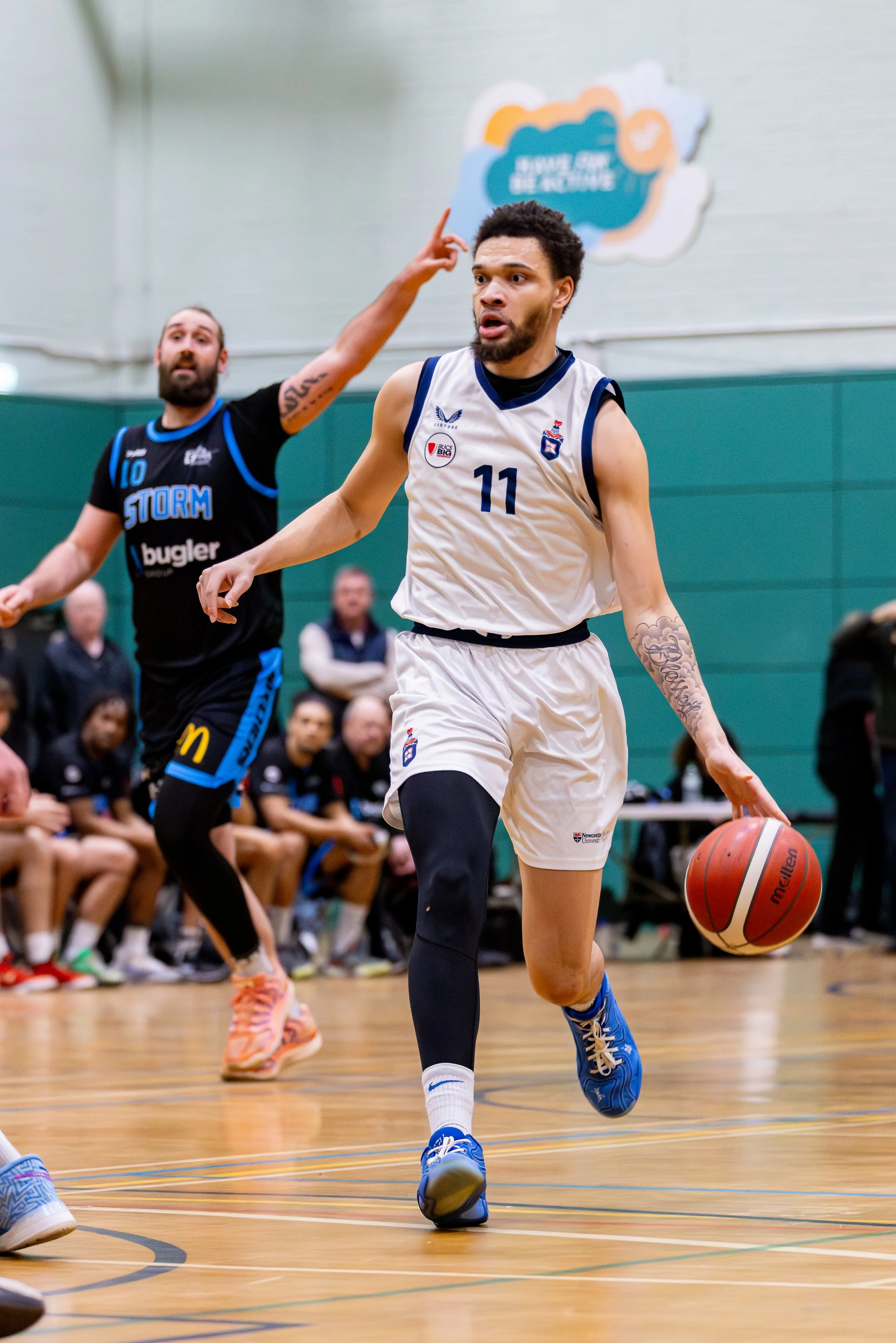 A basketball player wearing a white jersey with the number 11 dribbling a basketball on the court, while an opponent in a black jersey with the number 10 defends in the background. Spectators sit on the sidelines and watch the game.