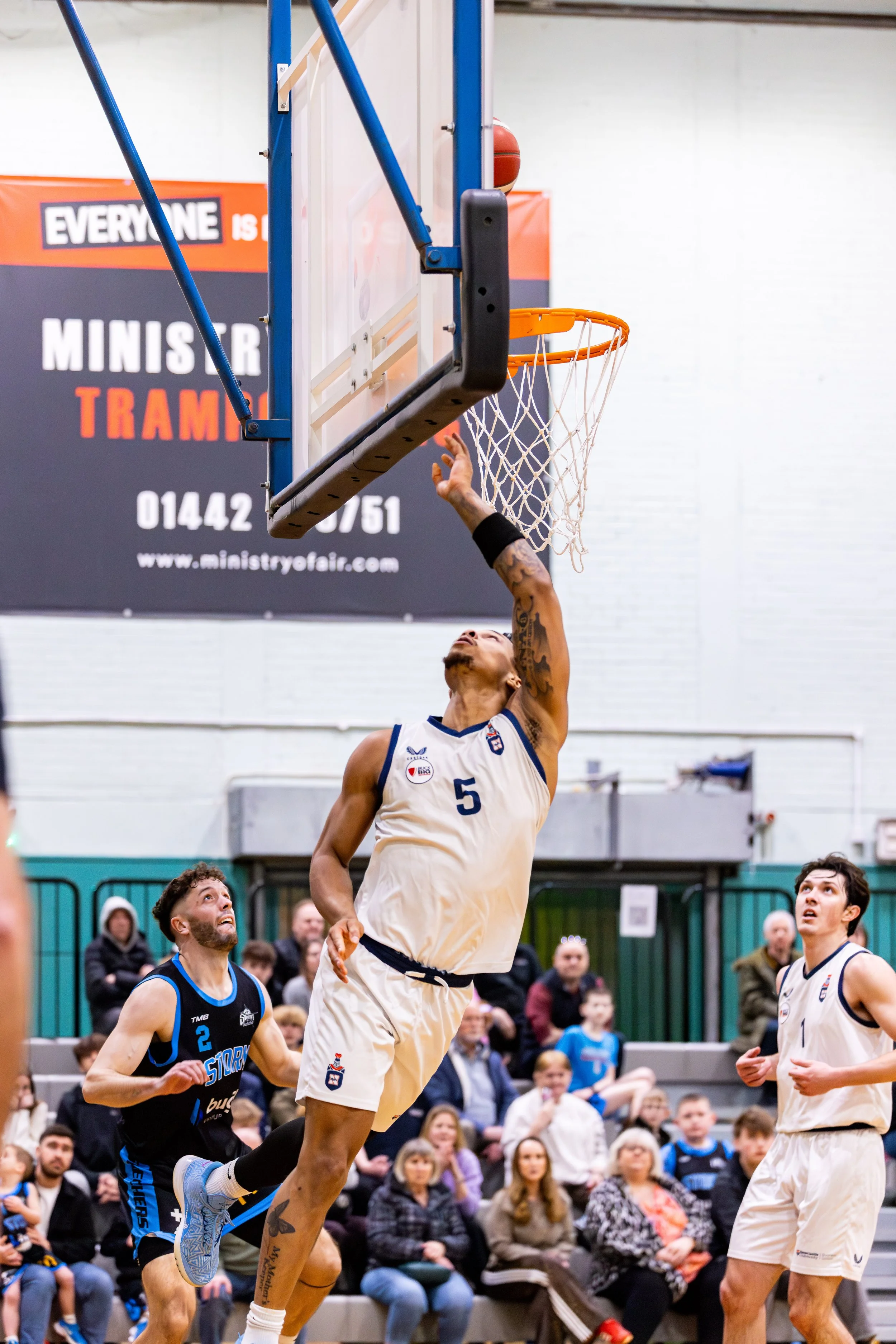 A basketball player in a white jersey jumps to make a shot near the basket, while a defender in a black jersey looks on and another teammate in a white jersey observes. The game is in progress in an indoor stadium with spectators in the background.