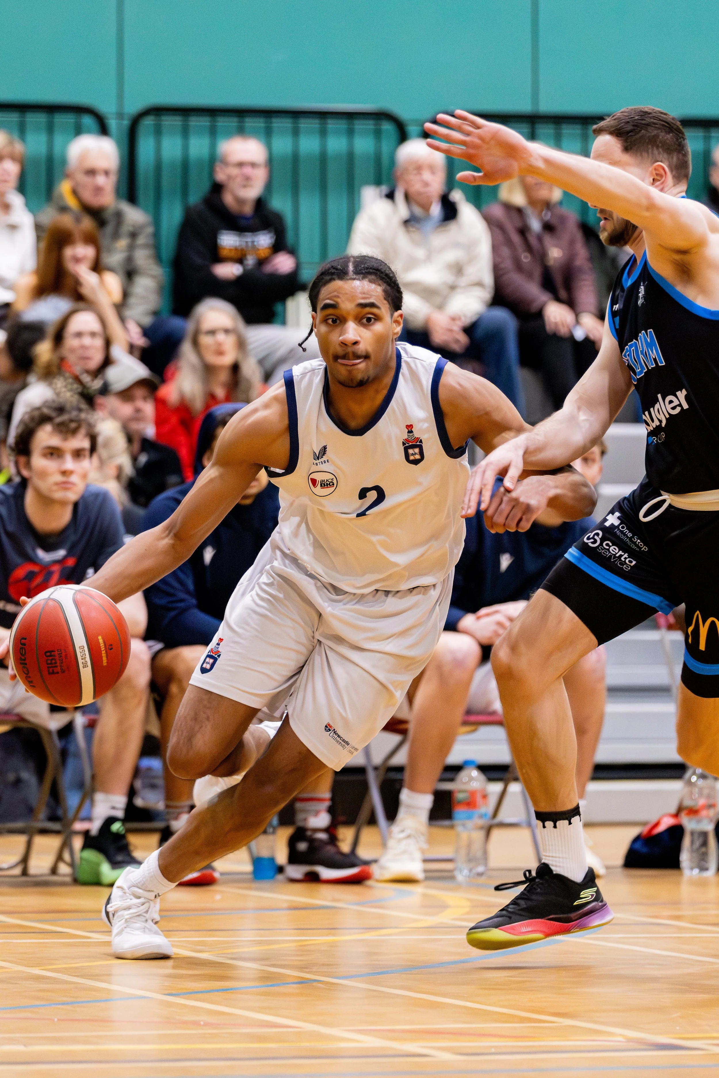 A basketball player in a white uniform dribbling the ball while being guarded by an opponent in a black uniform during a game with spectators watching from the bleachers.