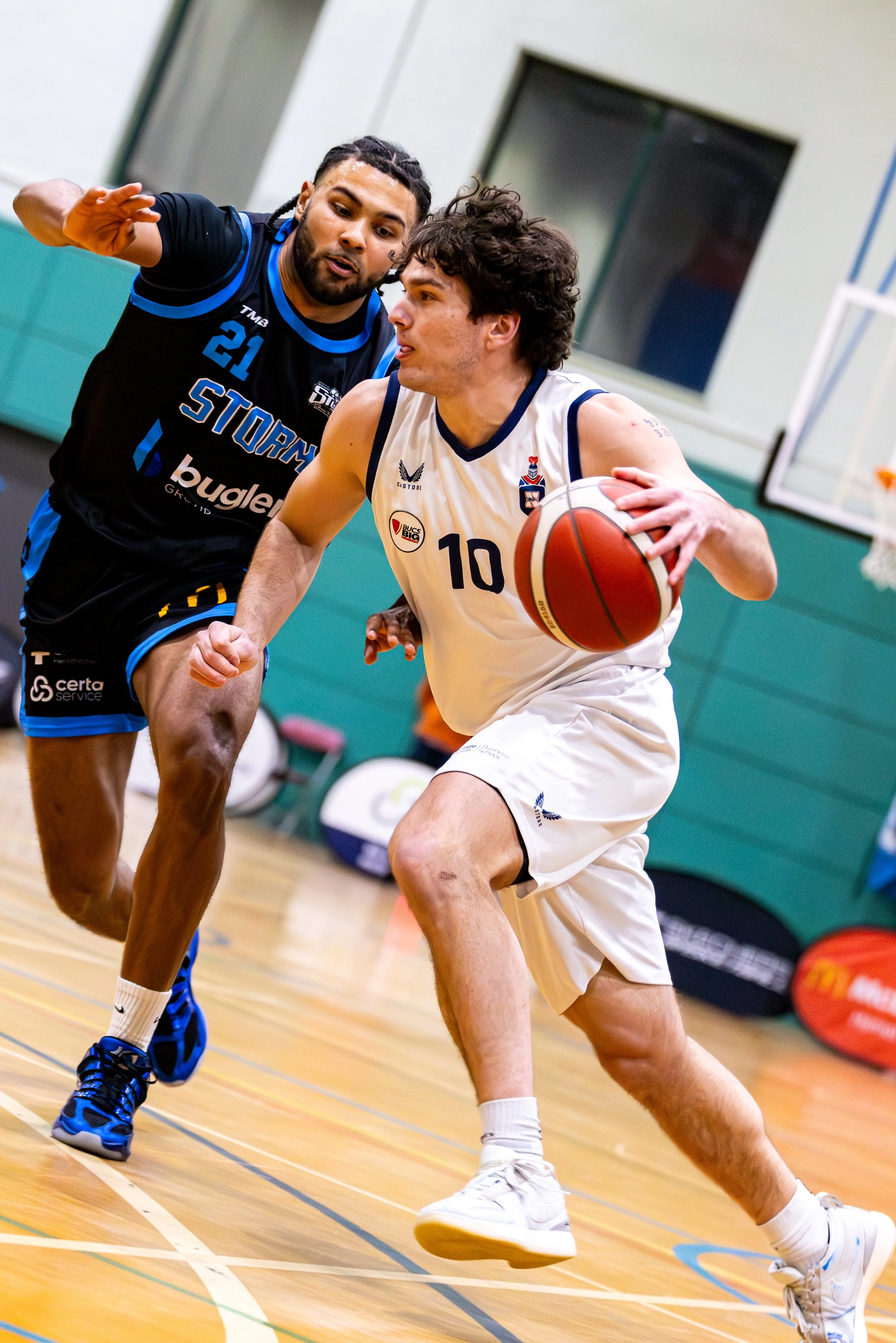Two basketball players competing for the ball during a game; one player in a white jersey with the number 10 and the other in a black jersey with blue accents and the number 21.