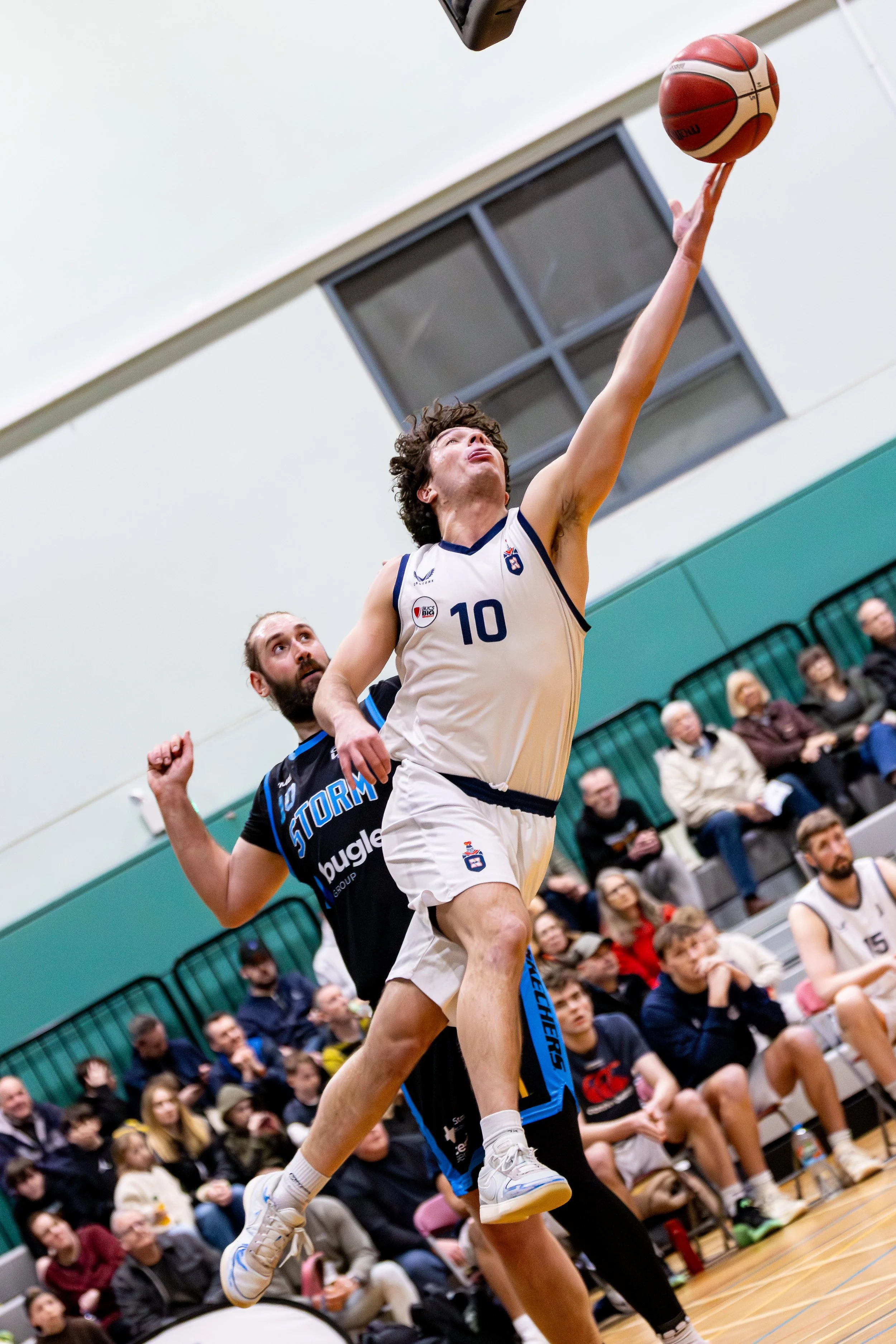 A basketball player wearing a white uniform with the number 10 is jumping to make a shot, while a defender in a black uniform attempts to block. The game is taking place indoors with spectators sitting on bleachers in the background.