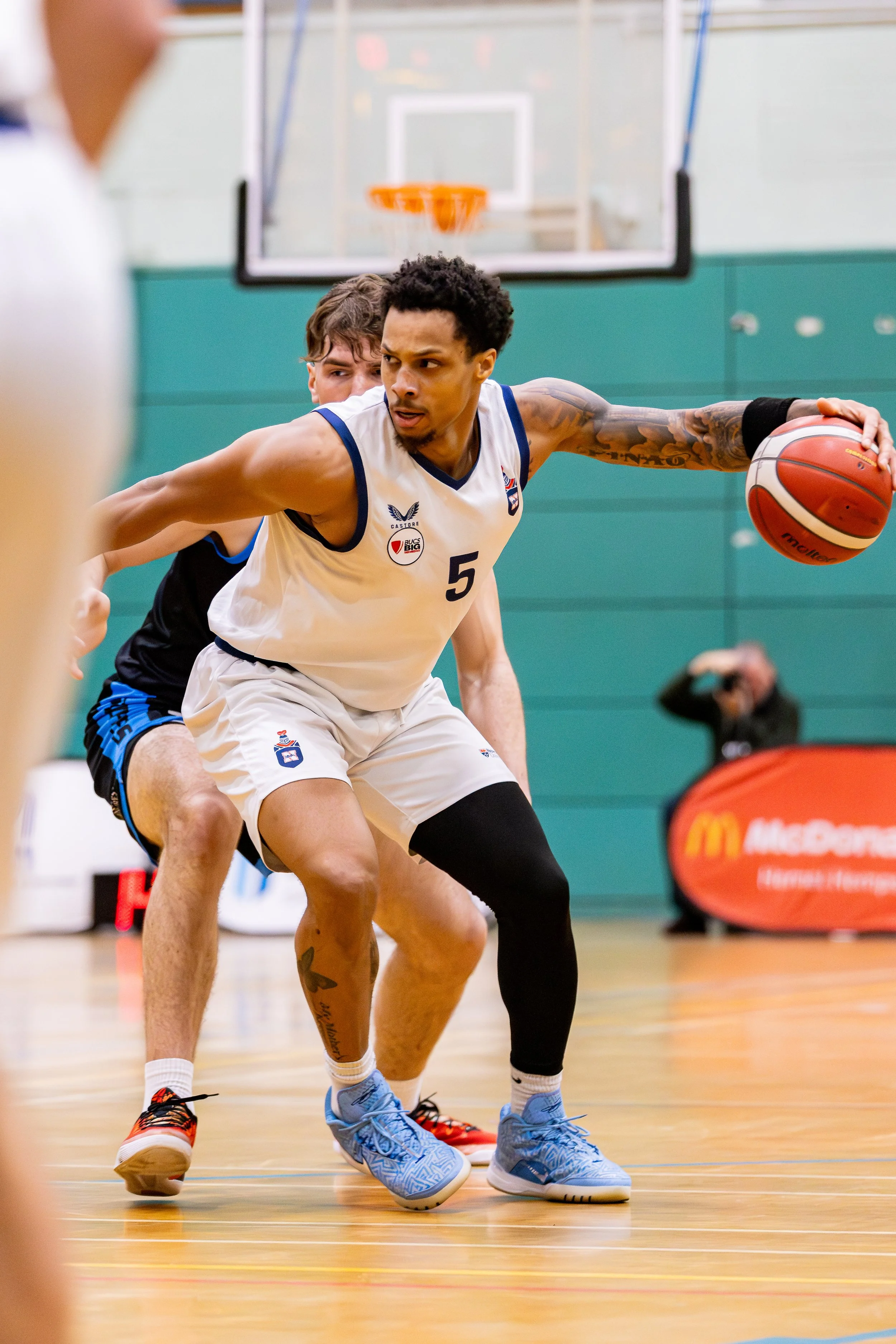 A basketball player in a white jersey with the number 5 dribbling the ball while being guarded closely by a defender in a black jersey in an indoor gymnasium.