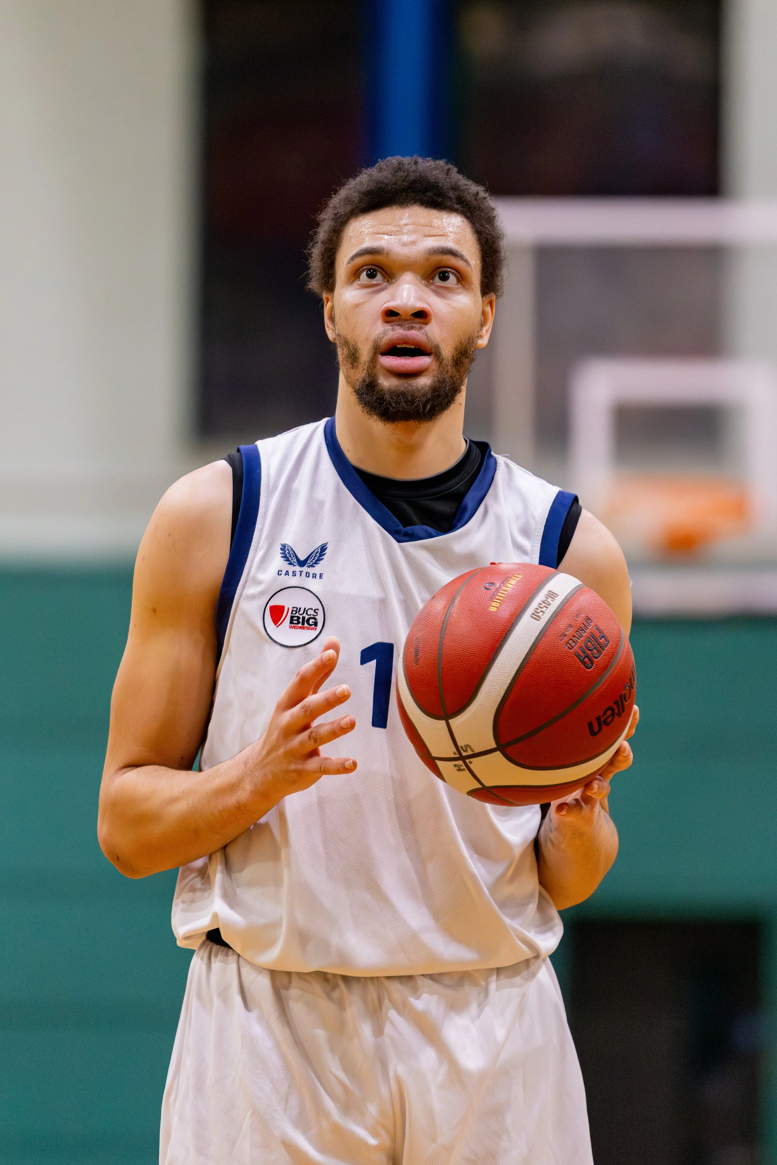 A basketball player preparing to shoot, holding a basketball with both hands, wearing a white jersey with blue accents and a black shirt underneath, standing on an indoor court.