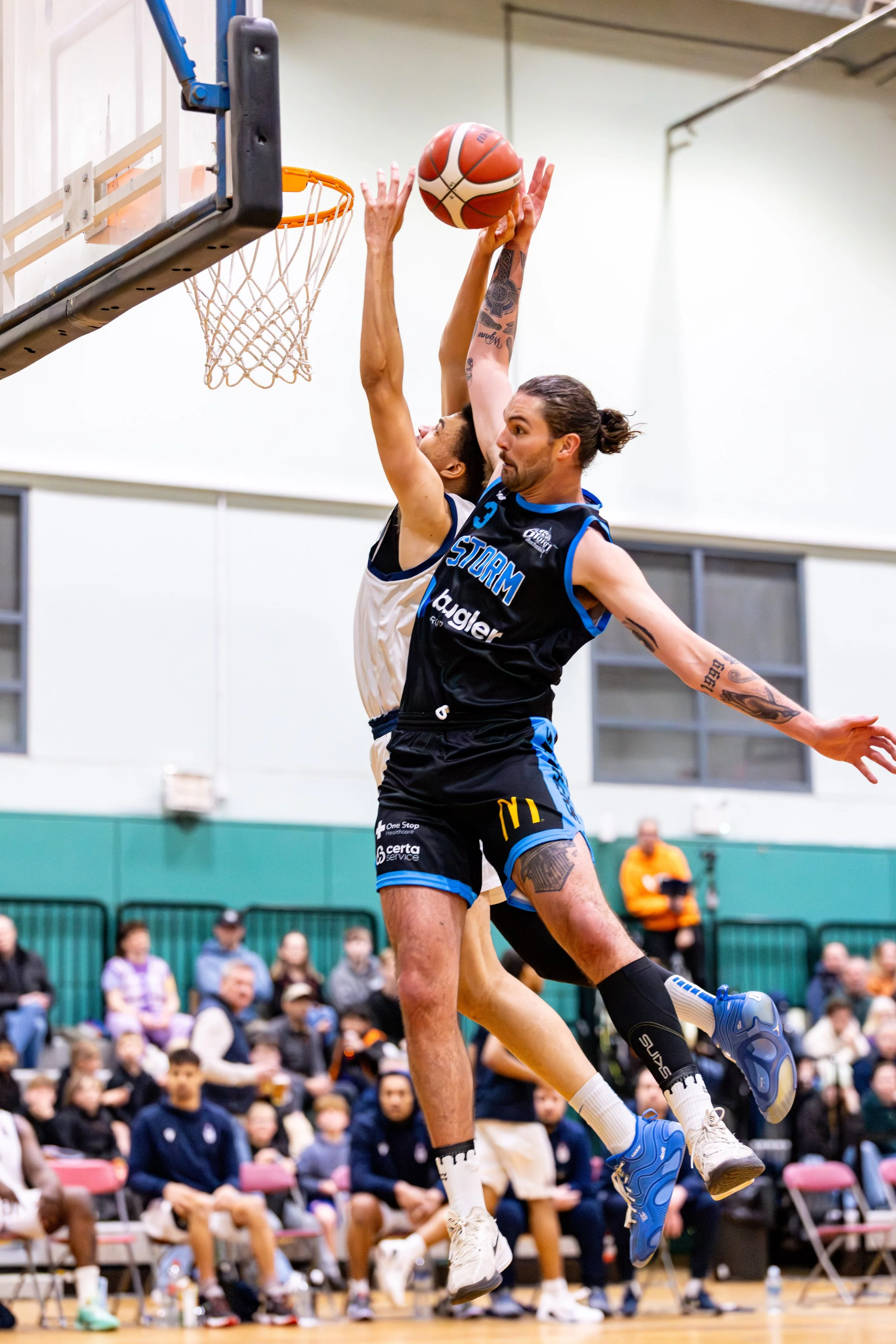 Two basketball players are jumping near the hoop, with one attempting a shot while the other tries to block it. The game is taking place in an indoor gym with spectators watching in the background.