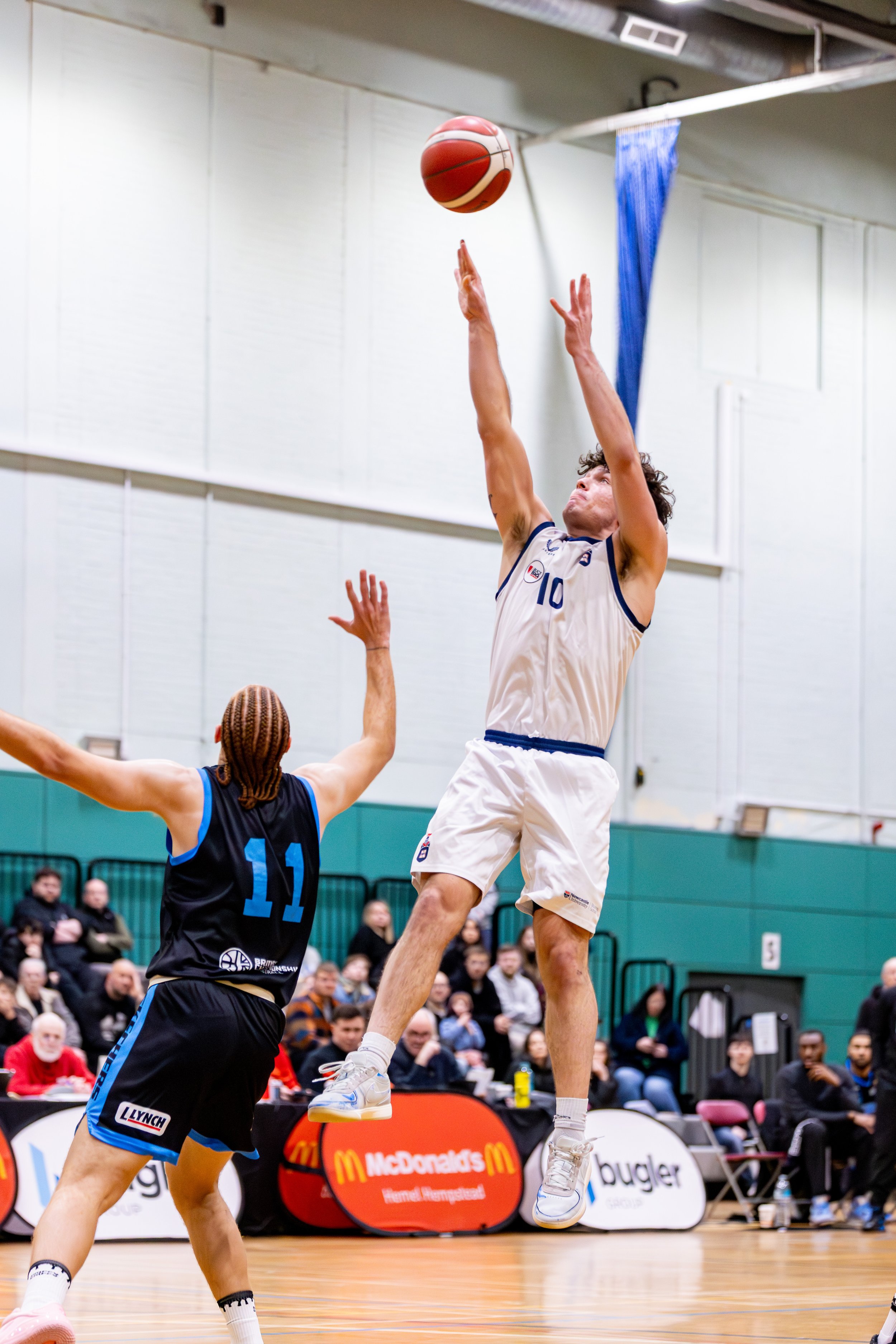 A basketball player in a white jersey with the number 10 jumps to shoot while an opponent in a navy jersey with the number 11 attempts to block. Spectators watch in the background.