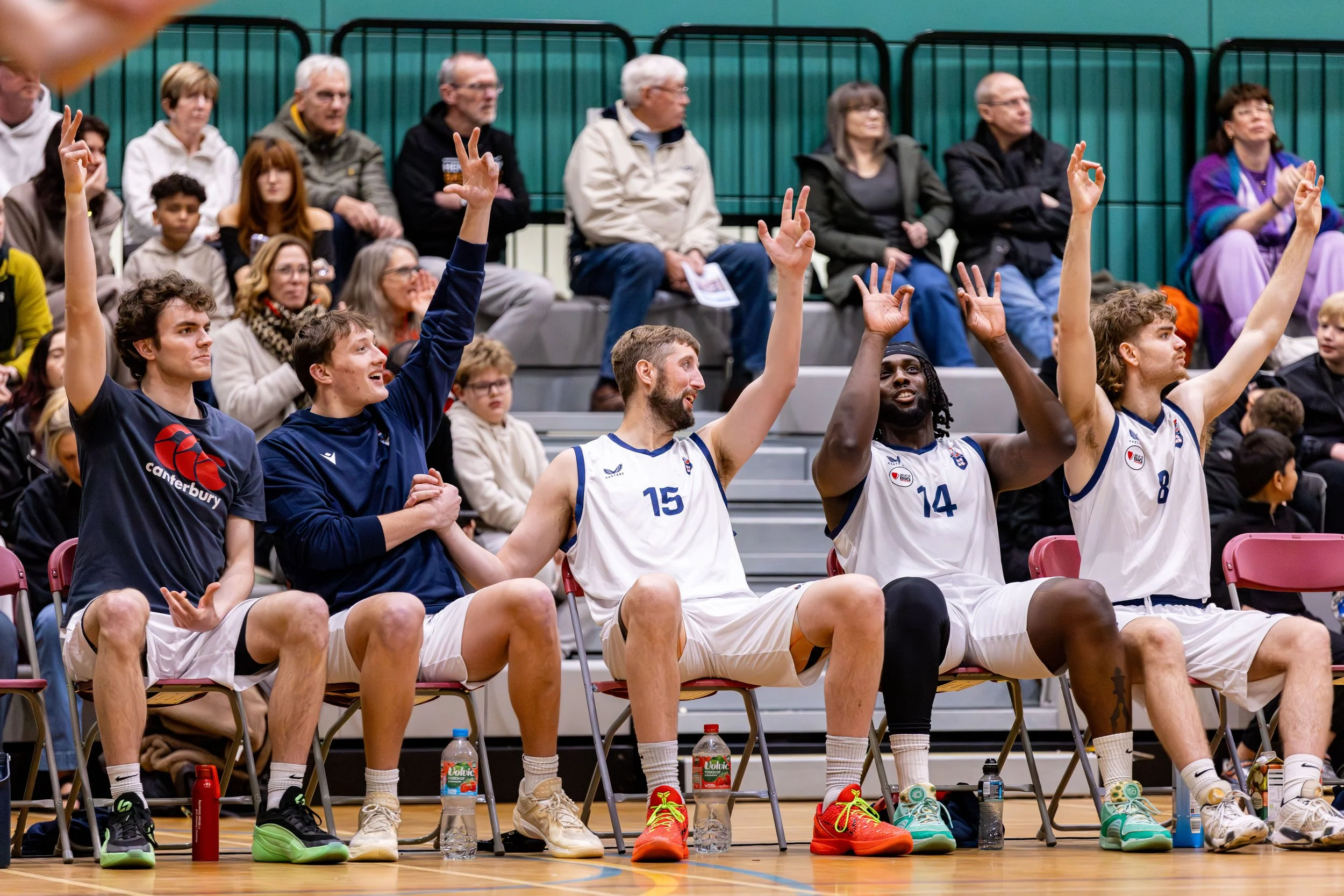 Basketball players sitting on chairs during a game, with some raising hands or making gestures, and spectators sitting behind them in the stands.
