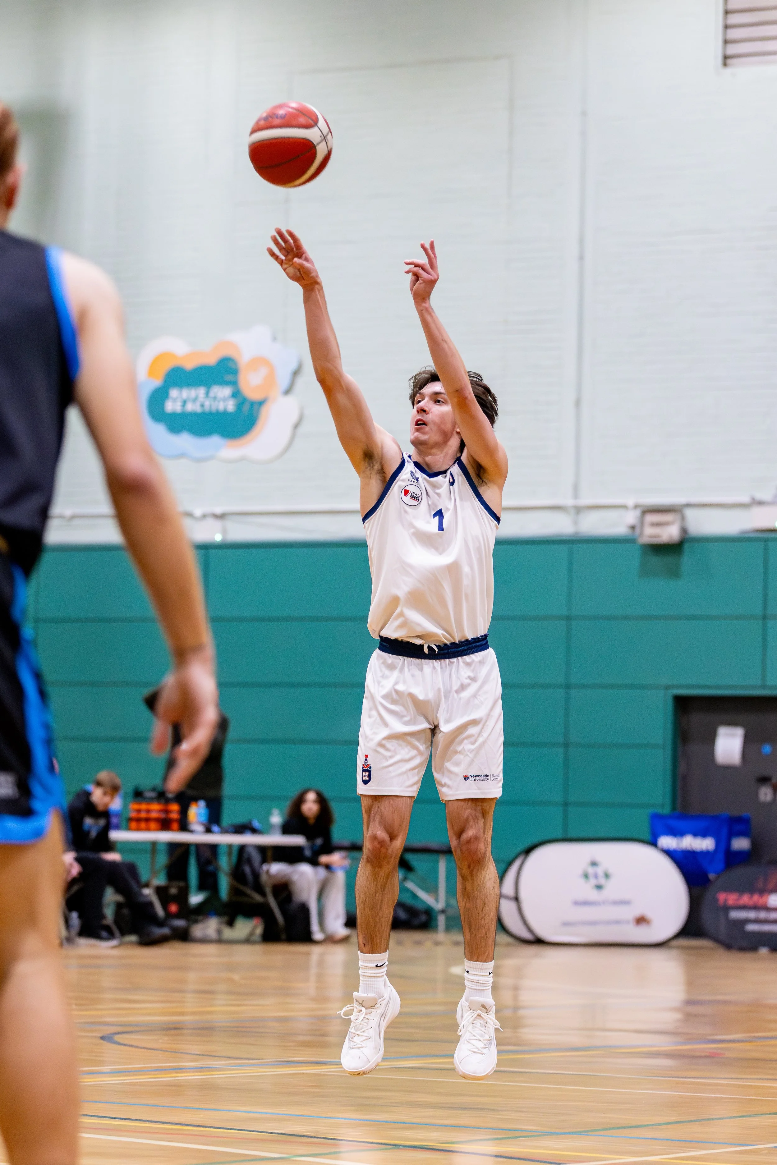 A young man wearing a white basketball uniform is jumping to shoot a basketball in an indoor gym. Other players and officials are visible in the background.