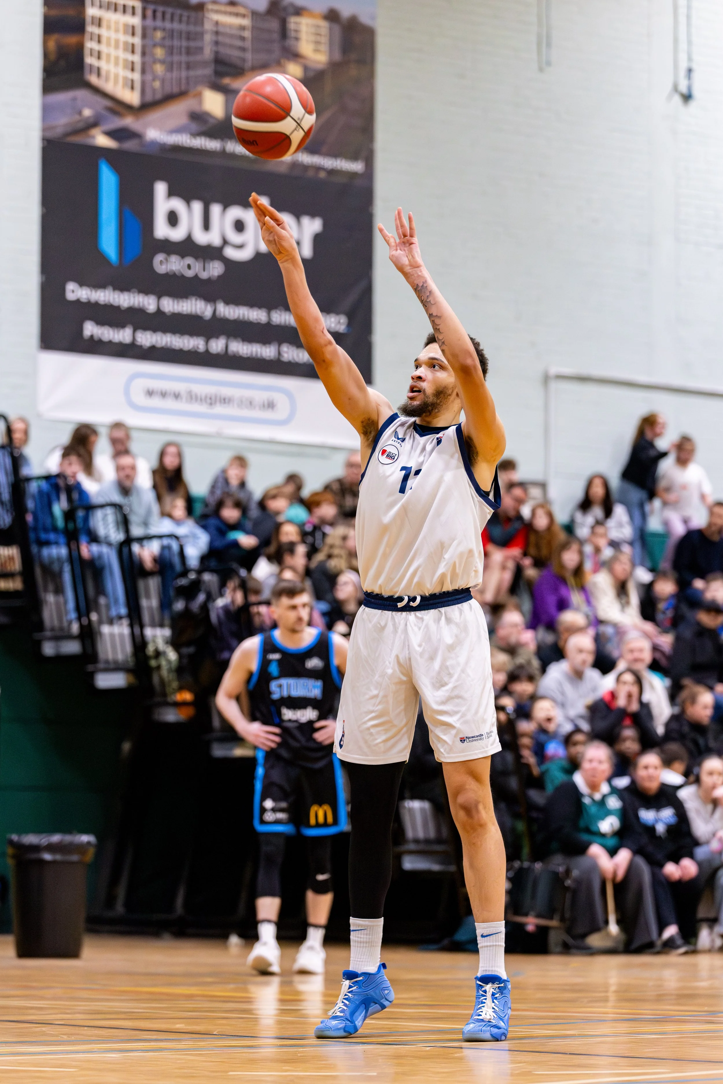 A basketball player in a white jersey and shorts, wearing a black sleeve on his left leg, is taking a shot during a game. The basketball is in mid-air above his hands. The gymnasium is filled with spectators and another player in a black and blue jer