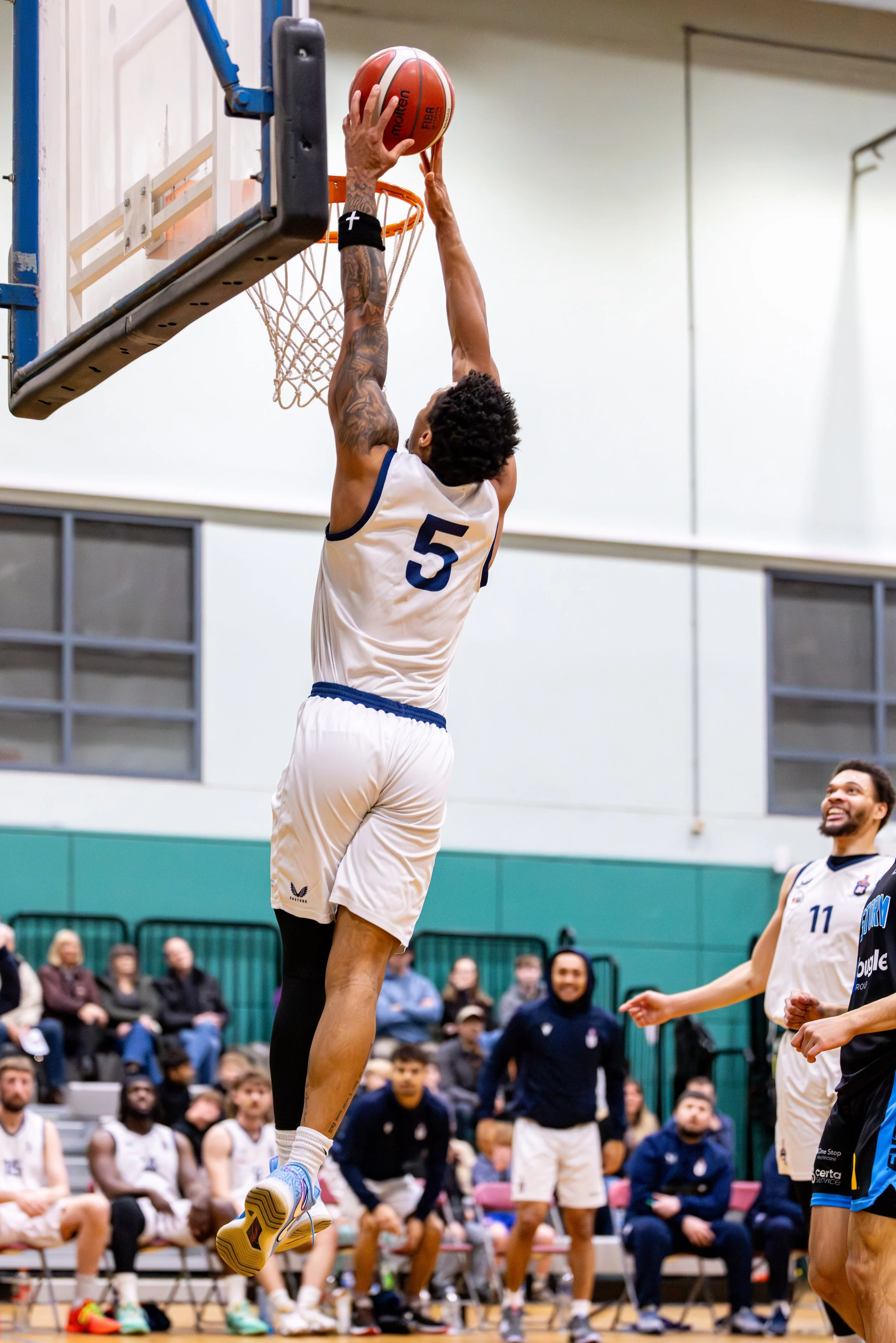 A basketball player wearing a white jersey with blue trim and the number 5 jumps high to dunk the ball into the hoop during a game, with spectators and teammates watching.