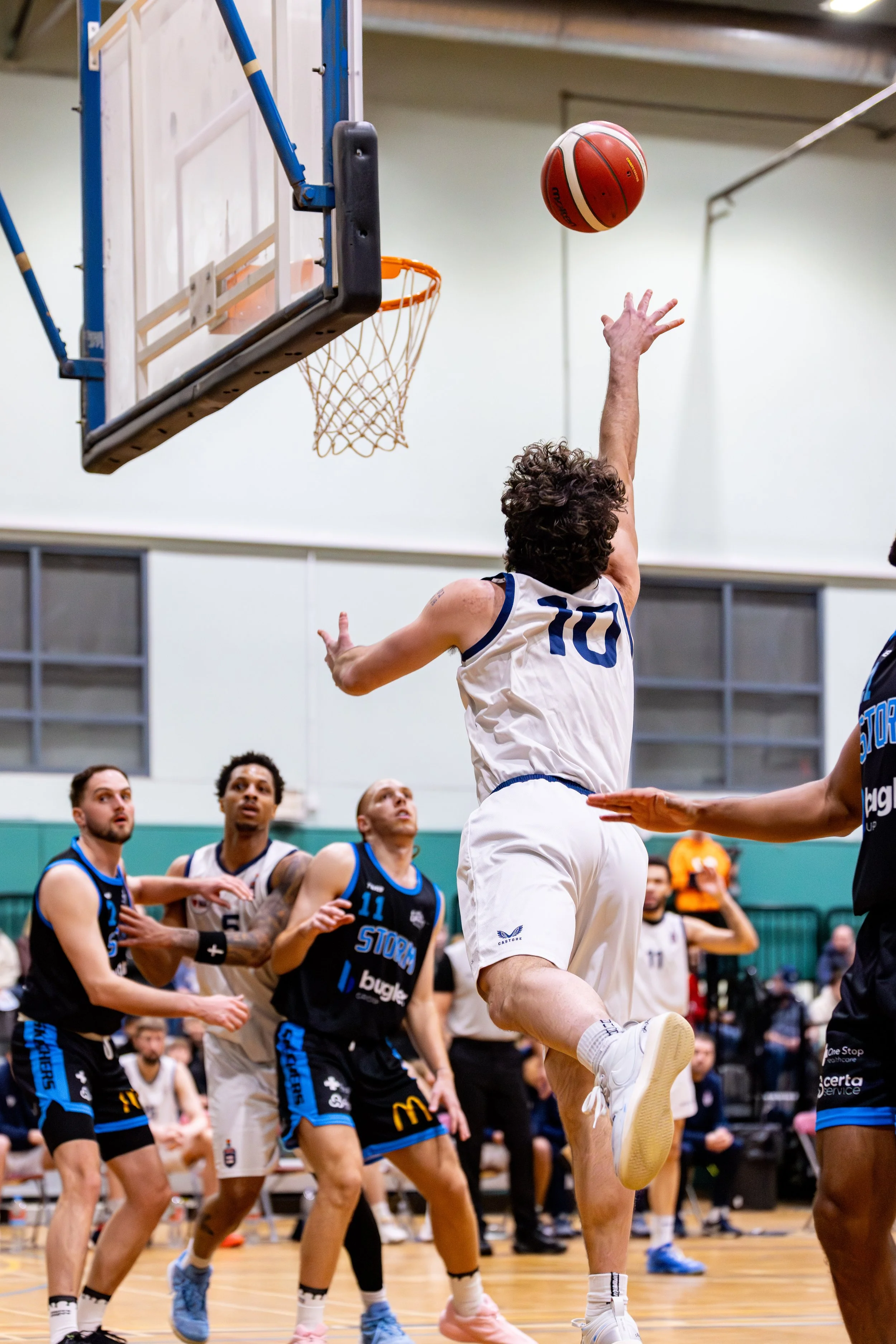 A basketball player wearing a white jersey with the number 10 is jumping towards the hoop for a layup shot during a game. Other players and spectators are visible in the background.