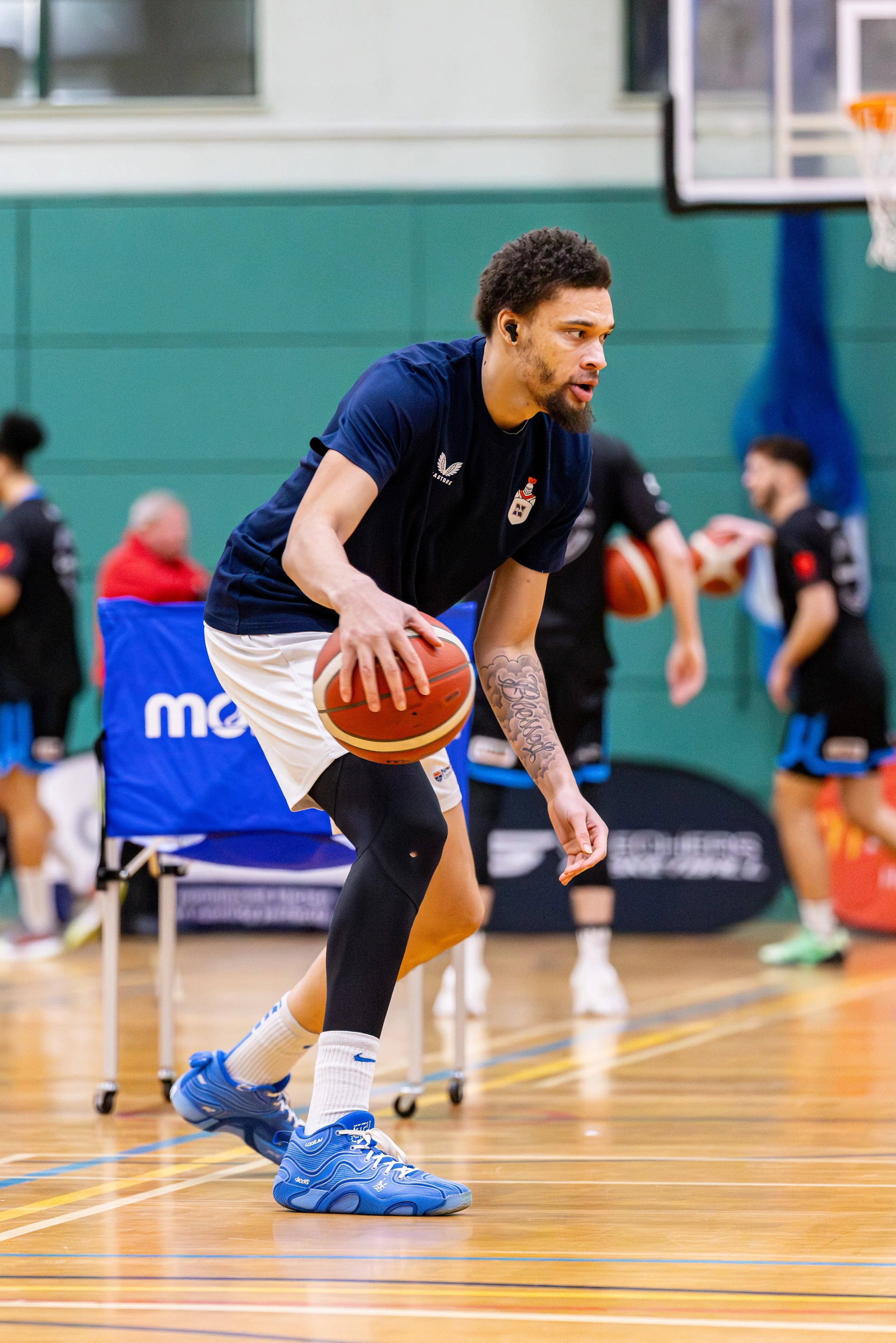 A man practicing basketball dribbling in a gymnasium, wearing a blue shirt, white shorts, black tights, and blue sneakers, with other players and basketball equipment in the background.