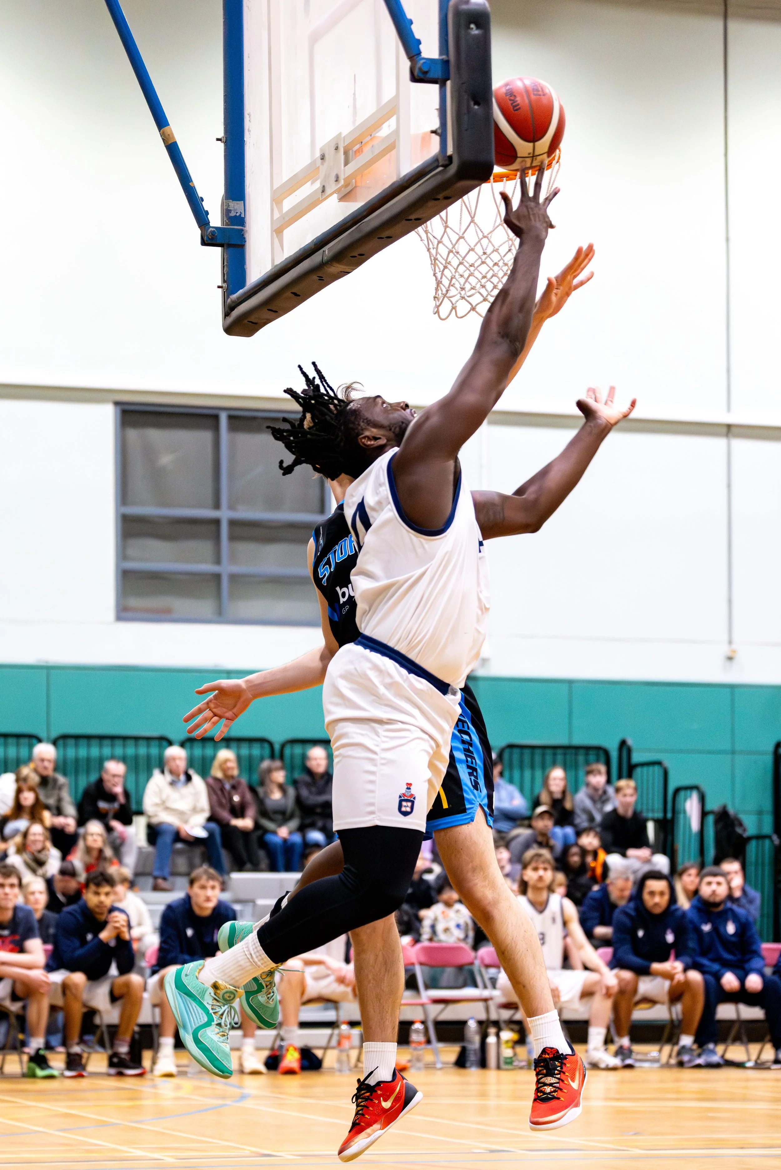 A basketball player is jumping to make a shot during a game, with spectators watching in the background.