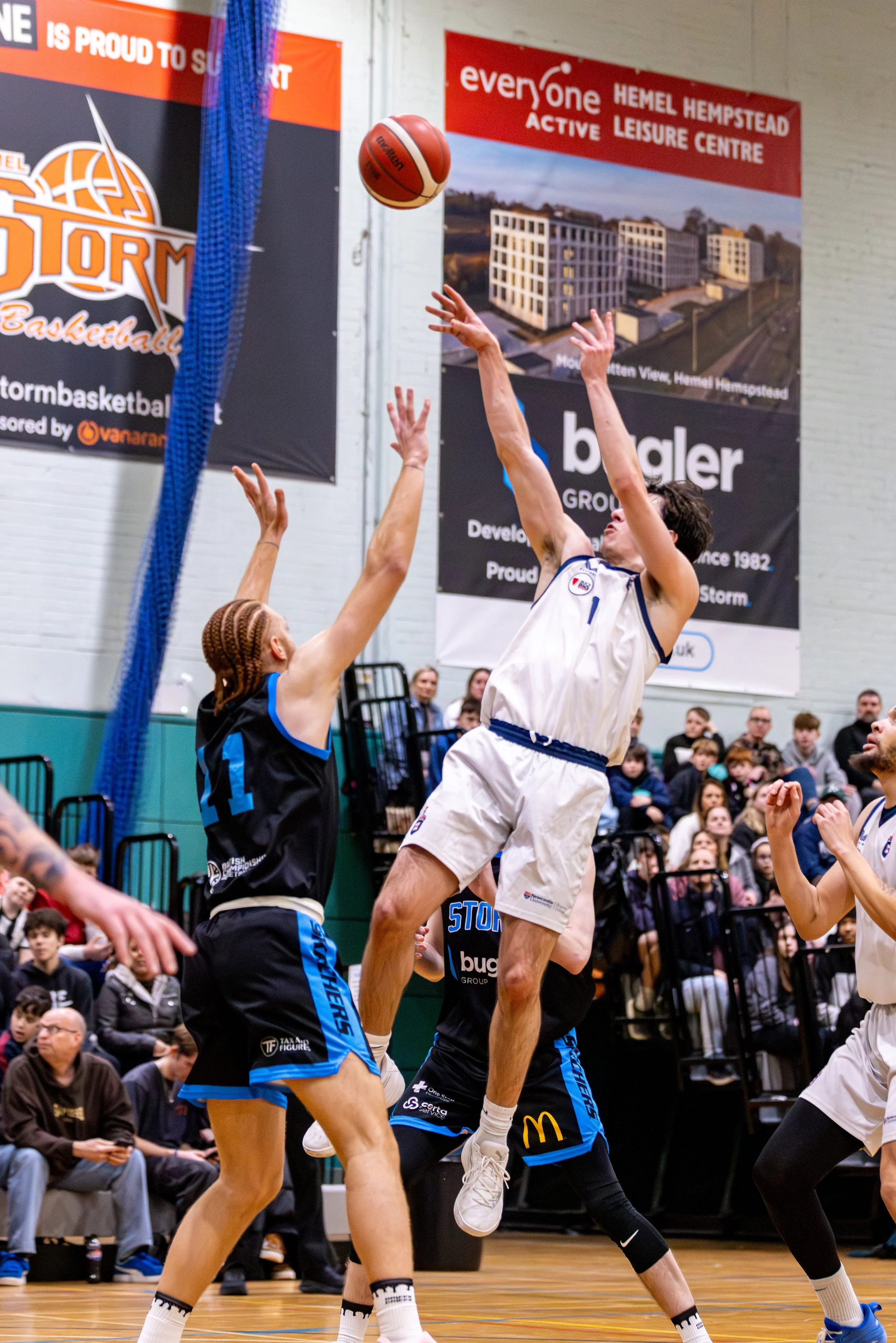 Two basketball players in action during a game, one in white jumping and shooting, the other in black and blue defending.