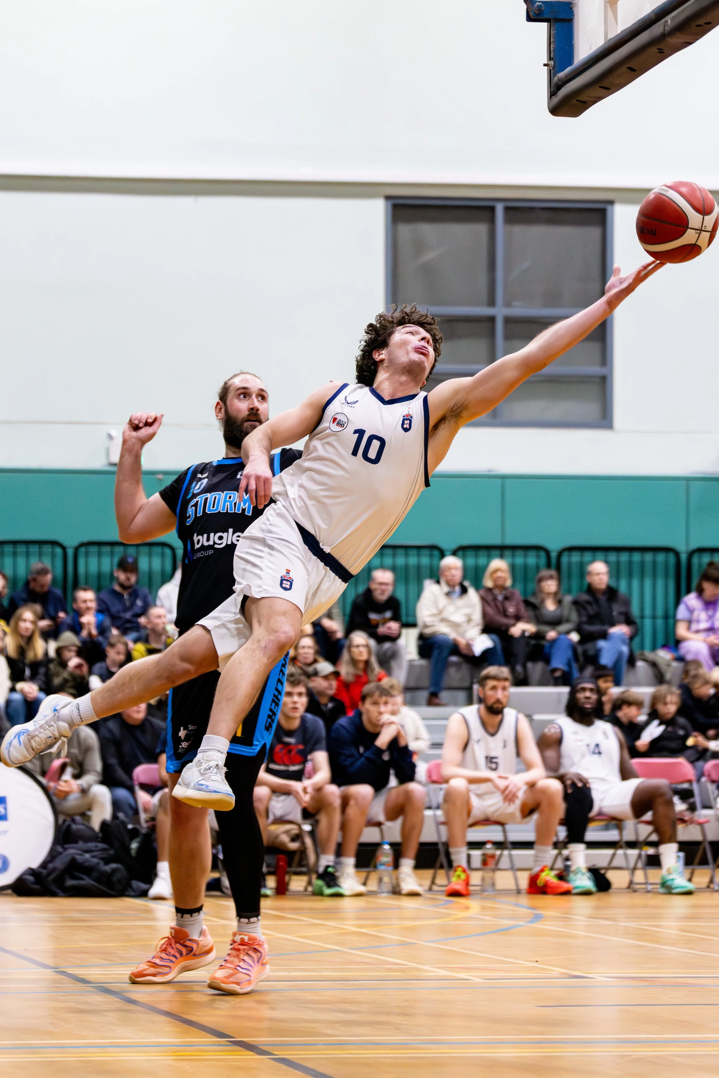 A basketball player in a white jersey with the number 10 reaches for the basket during a game, while another player in a black jersey attempts to defend. The game is indoor with spectators in the background.