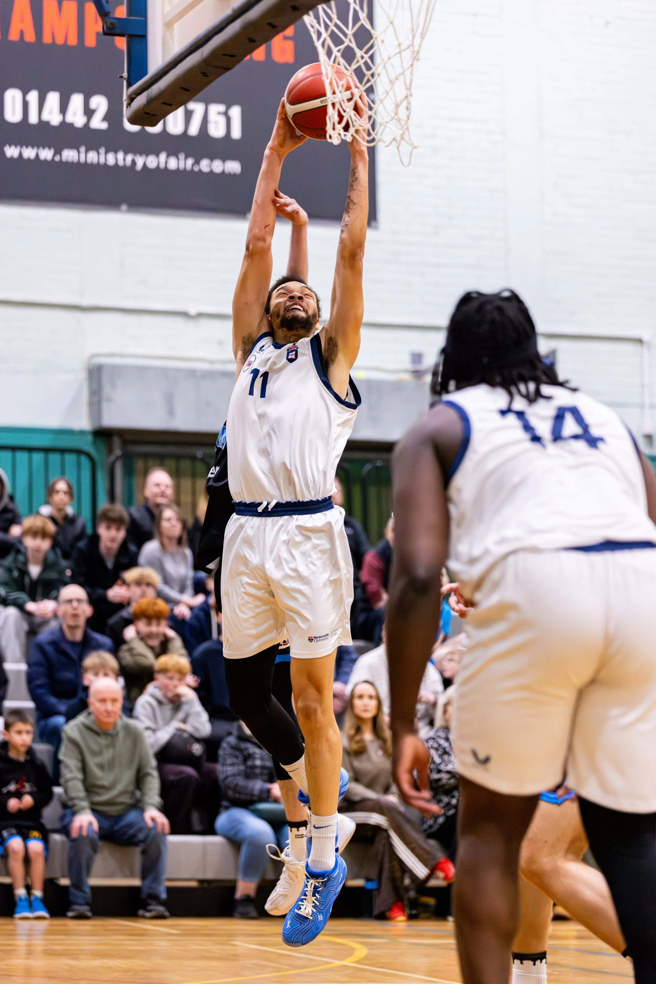 A basketball player wearing a white jersey with the number 11 jumping to make a shot at the basket while another player watches.
