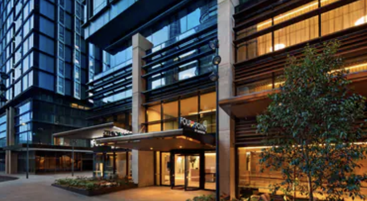 Modern multi-story building with glass windows and a lit entrance, featuring a small garden and a tree outside.