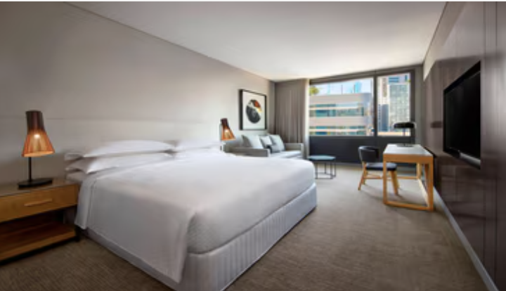 Hotel room with a large bed, bedside tables with lamps, a sofa, a work desk, and a flat-screen TV, with a window showing city buildings.