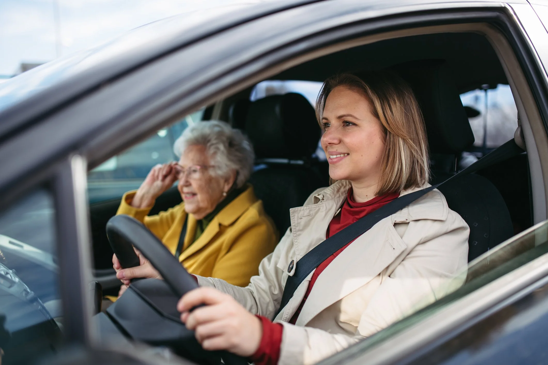 A young woman driving a car with an elderly woman sitting in the passenger seat, both smiling.