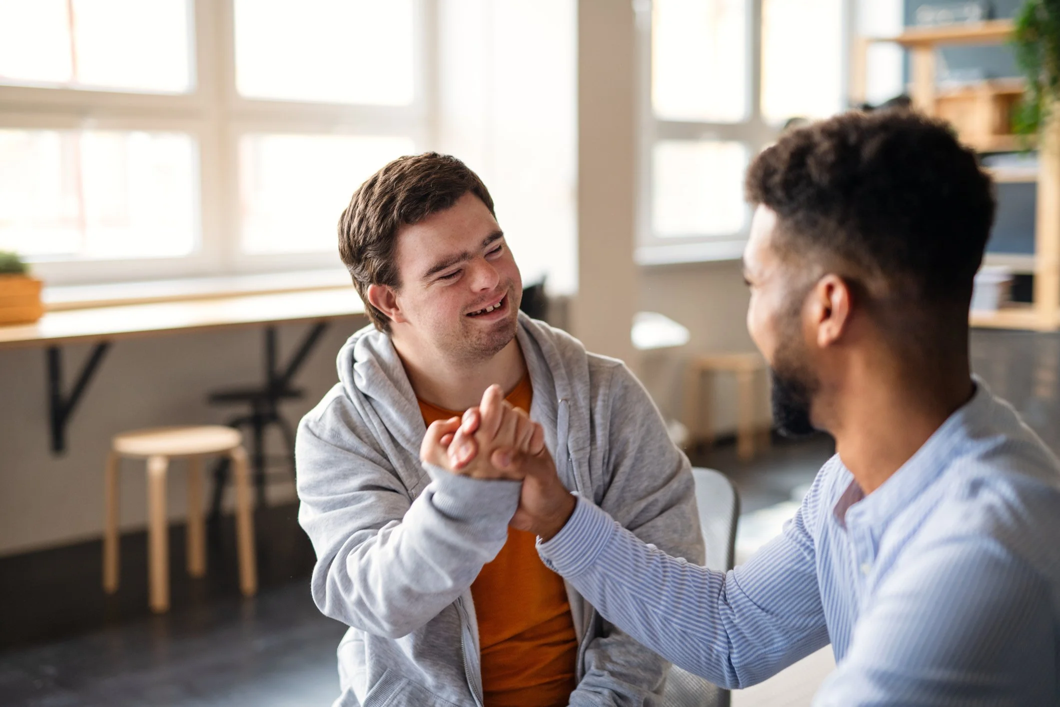 Two men are doing a fist bump and smiling at each other in a bright room with large windows.