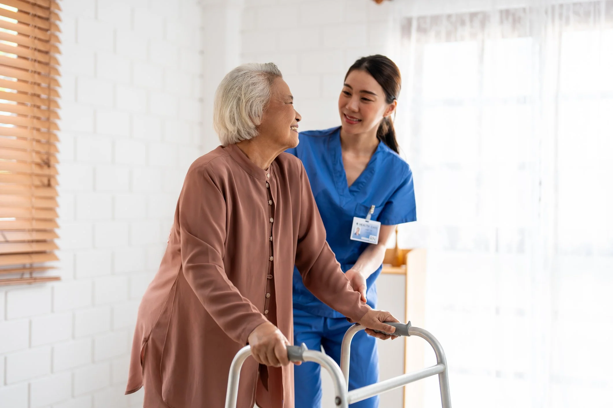 Elderly woman using a walker assisted by a healthcare worker in blue scrubs.