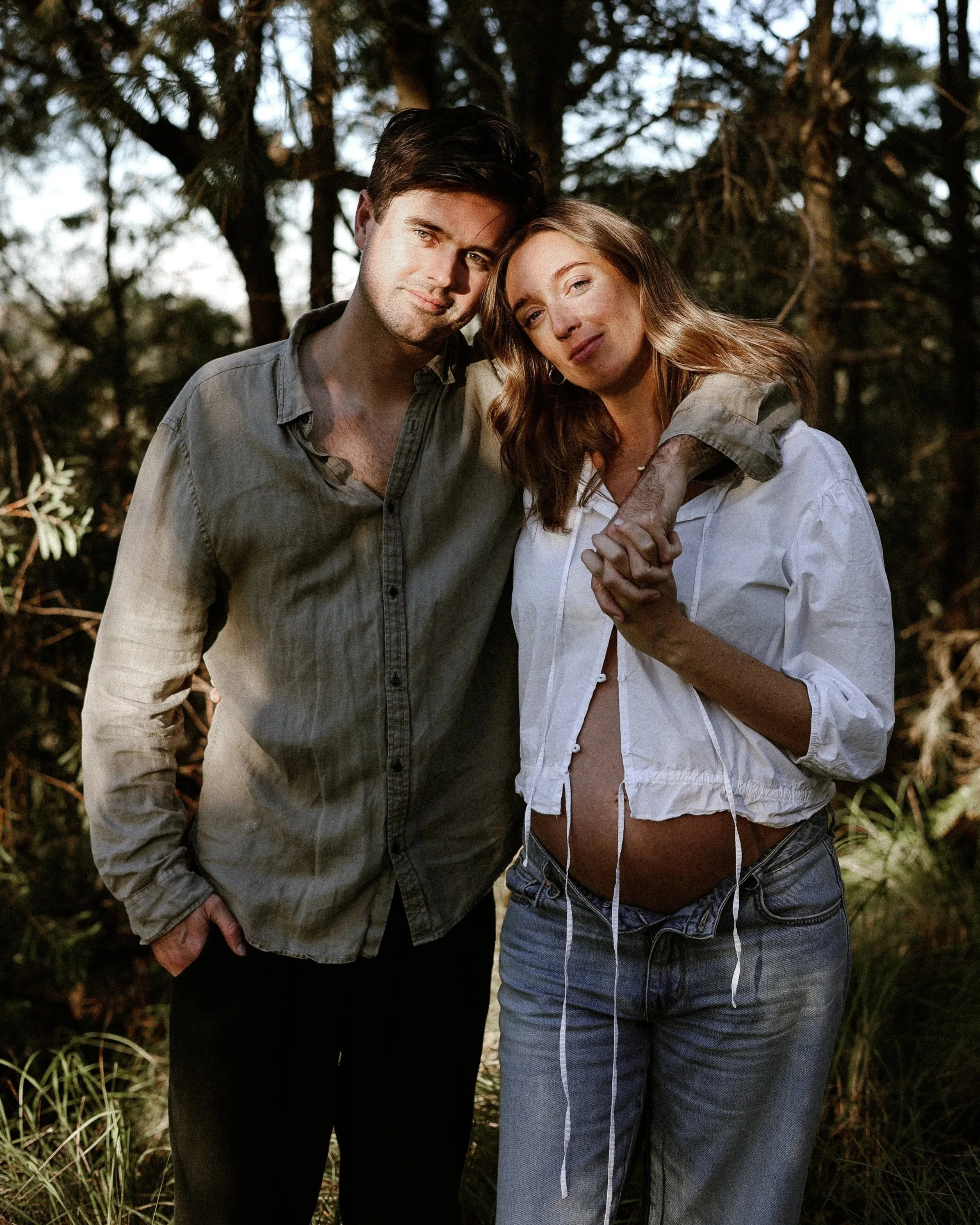 A young man and woman (who is 8 months pregnant) standing close together outdoors, with their heads touching and smiling at the camera, surrounded by trees and greenery. This is a maternity photo shoot.