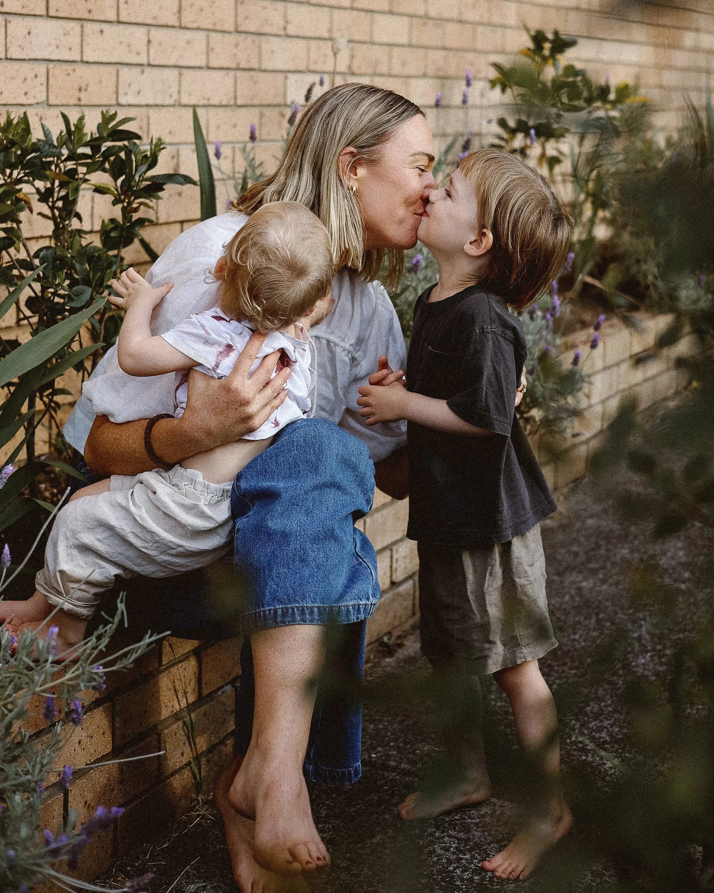 A woman with two young children enjoying a moment together outside near a brick wall with plants and purple flowers. The oldest son gives his mum a kiss.