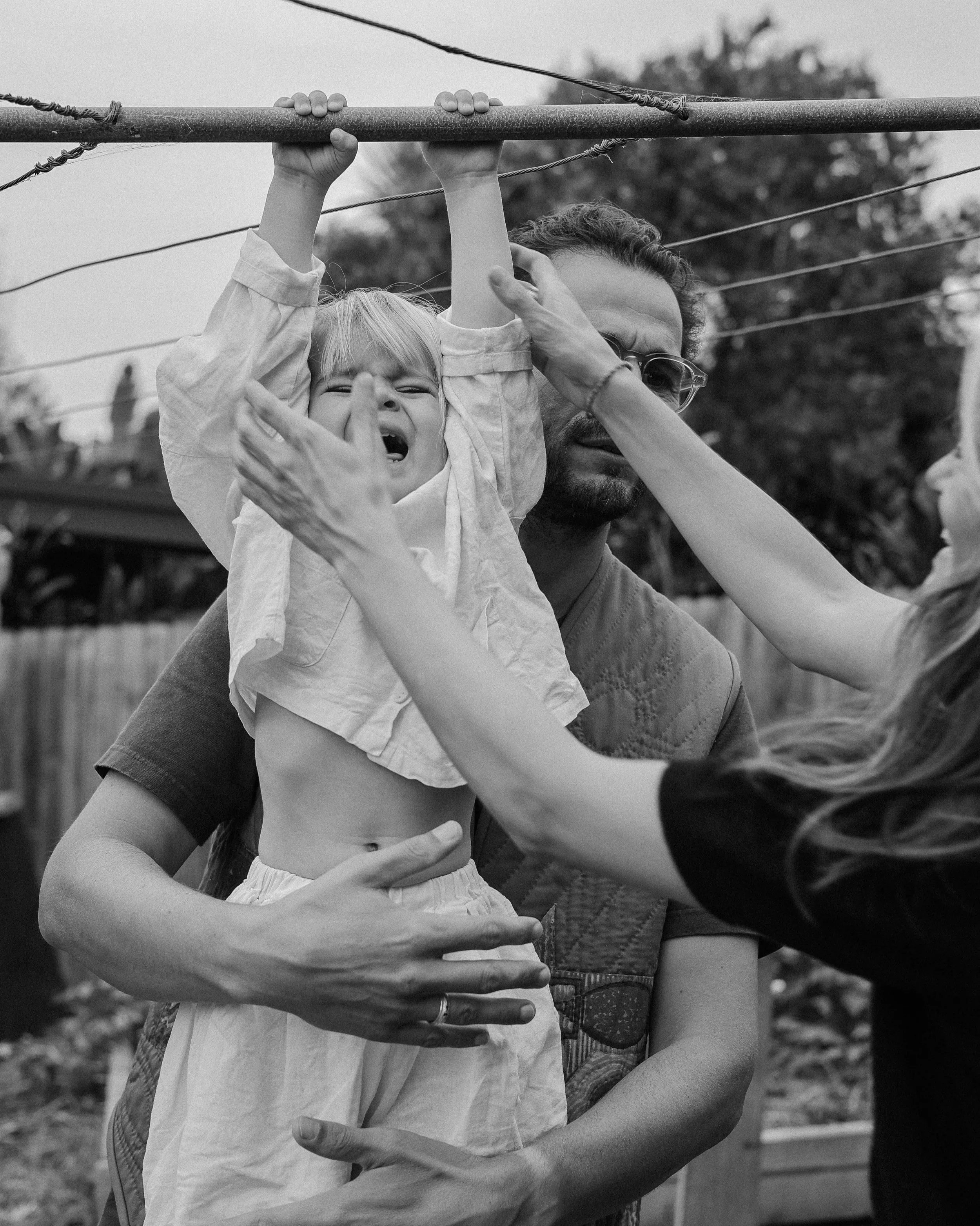 A young  hanging from a washing line. The boy appears to be crying, with his eyes closed, while the dad is holding him around the waist. The image is in black and white, and the background shows trees and a fence.