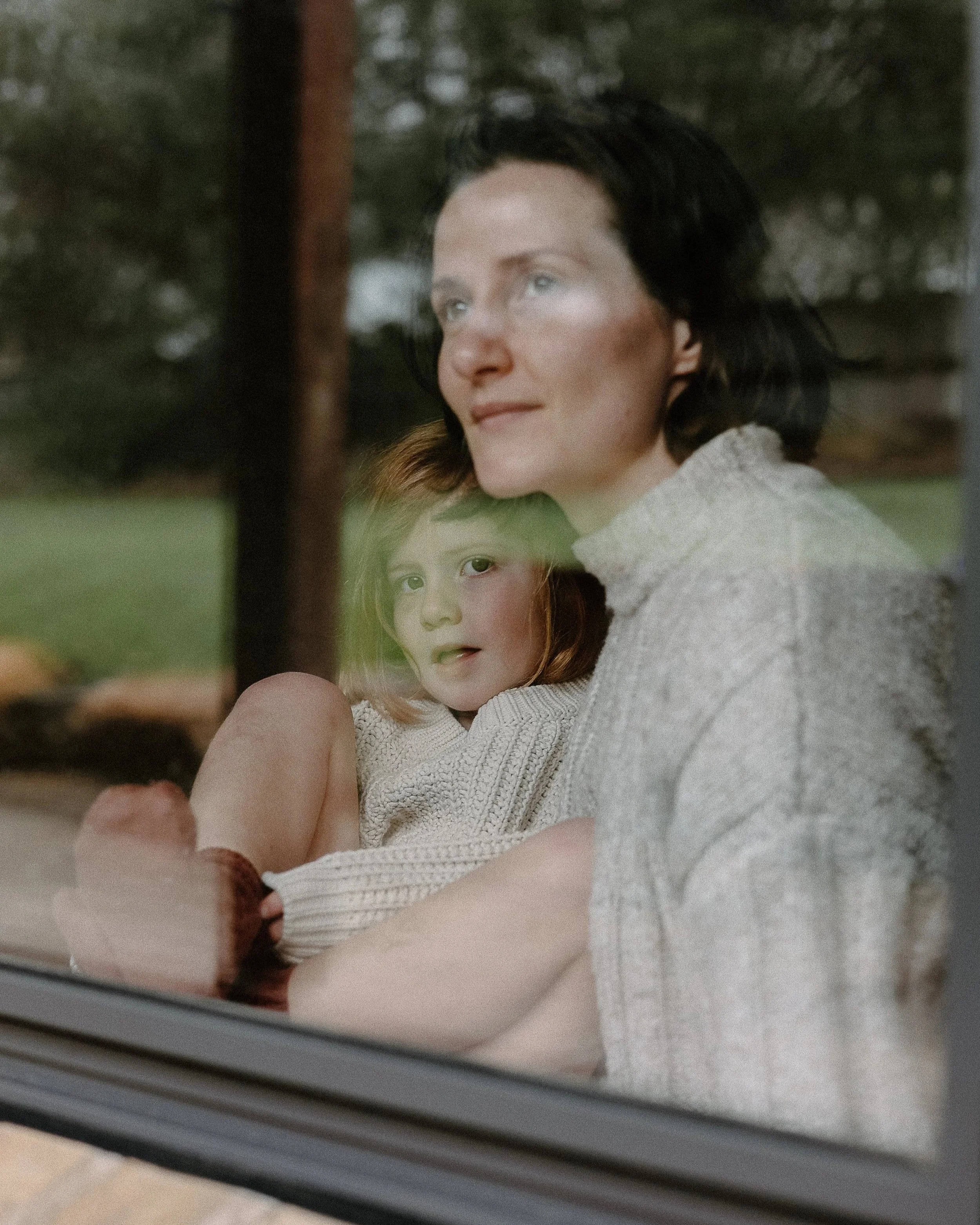 Reflection of a woman with dark hair and blue eyes, and a young girl with red hair and fair skin, photographed through a window. The woman is wearing a light-colored sweater and the girl has a curious expression.