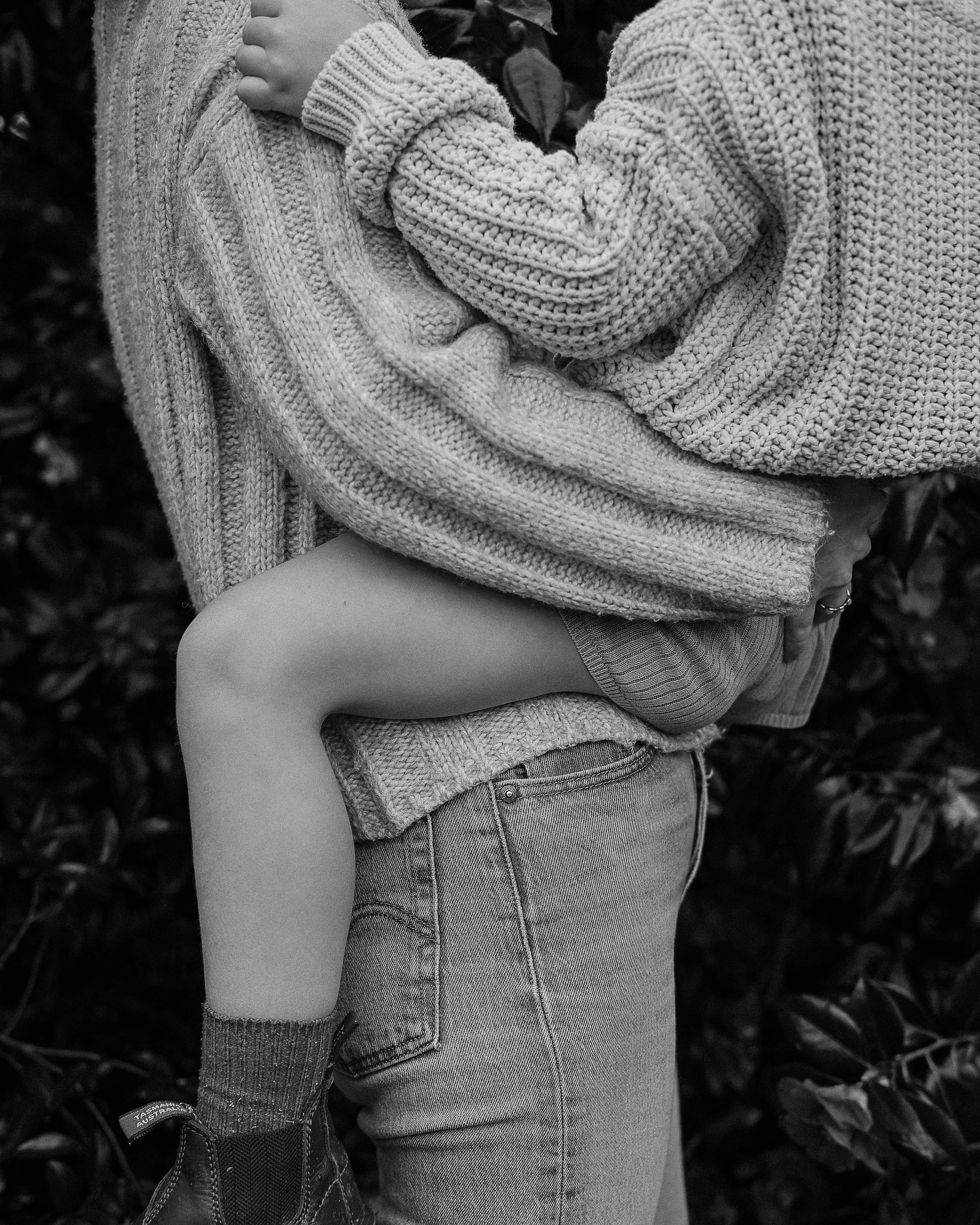 Close-up of a child wearing a chunky knit sweater with a textured pattern, holding onto an adult's sweater. The child is dressed in casual denim shorts, and the adult is wearing a patterned skirt or dress. The background shows blurred foliage, indica