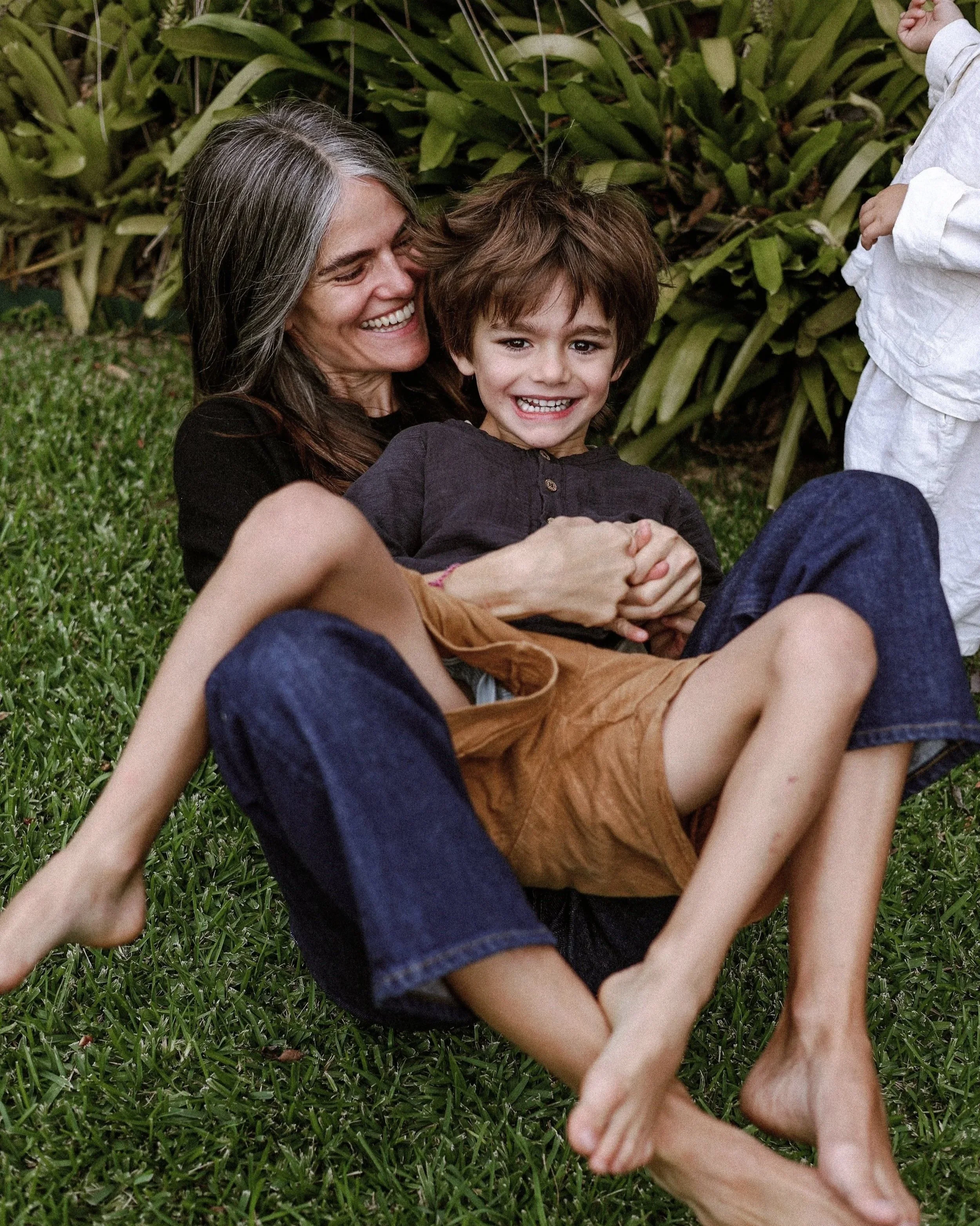 An young mother and her young boy lying on green grass, smiling and embracing, with leafy bushes in the background.