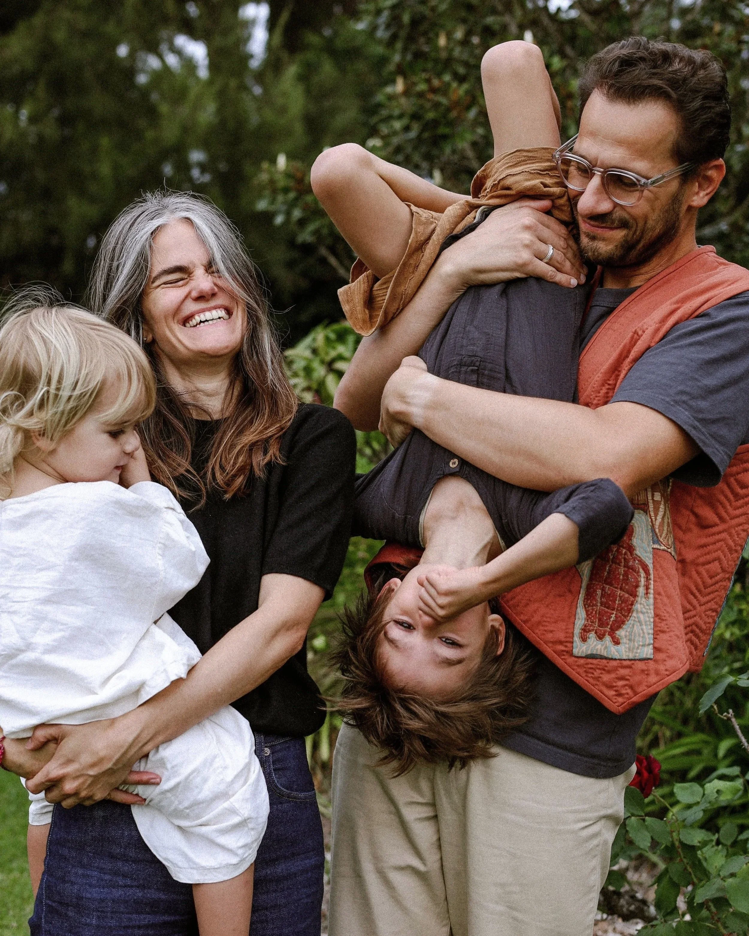 Two children and two adults smiling and interacting outdoors among greenery, with one of the children being lifted upside down and the others observing for a family portrait session on the South Coast