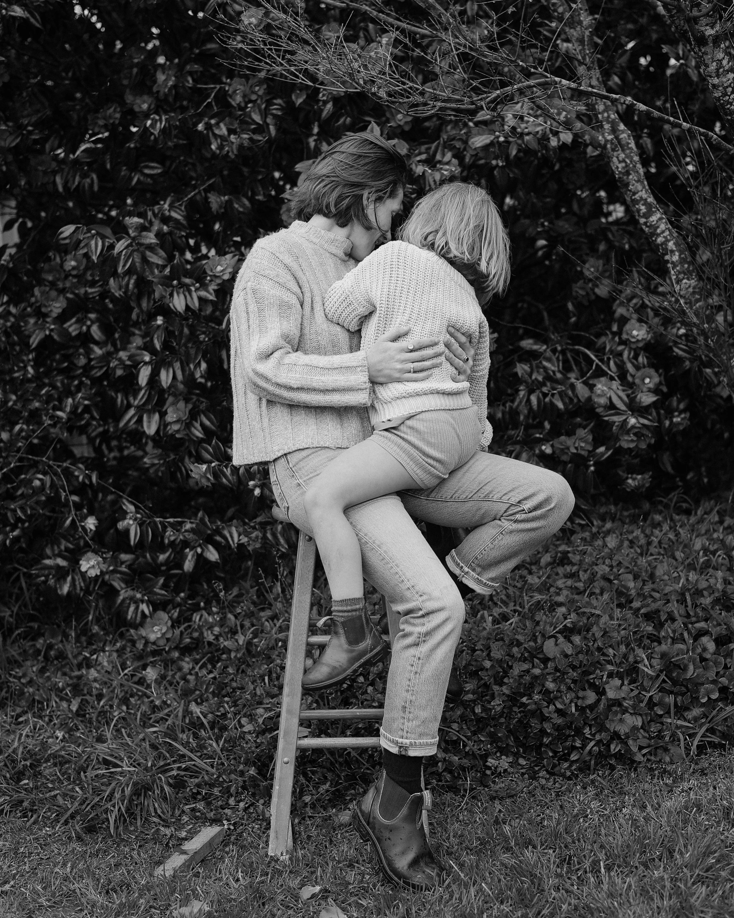 A person sitting on a stool wearing a sweater and boots, holding a child dressed in a sweater, with a background of dense shrubbery and tree branches, in black and white.