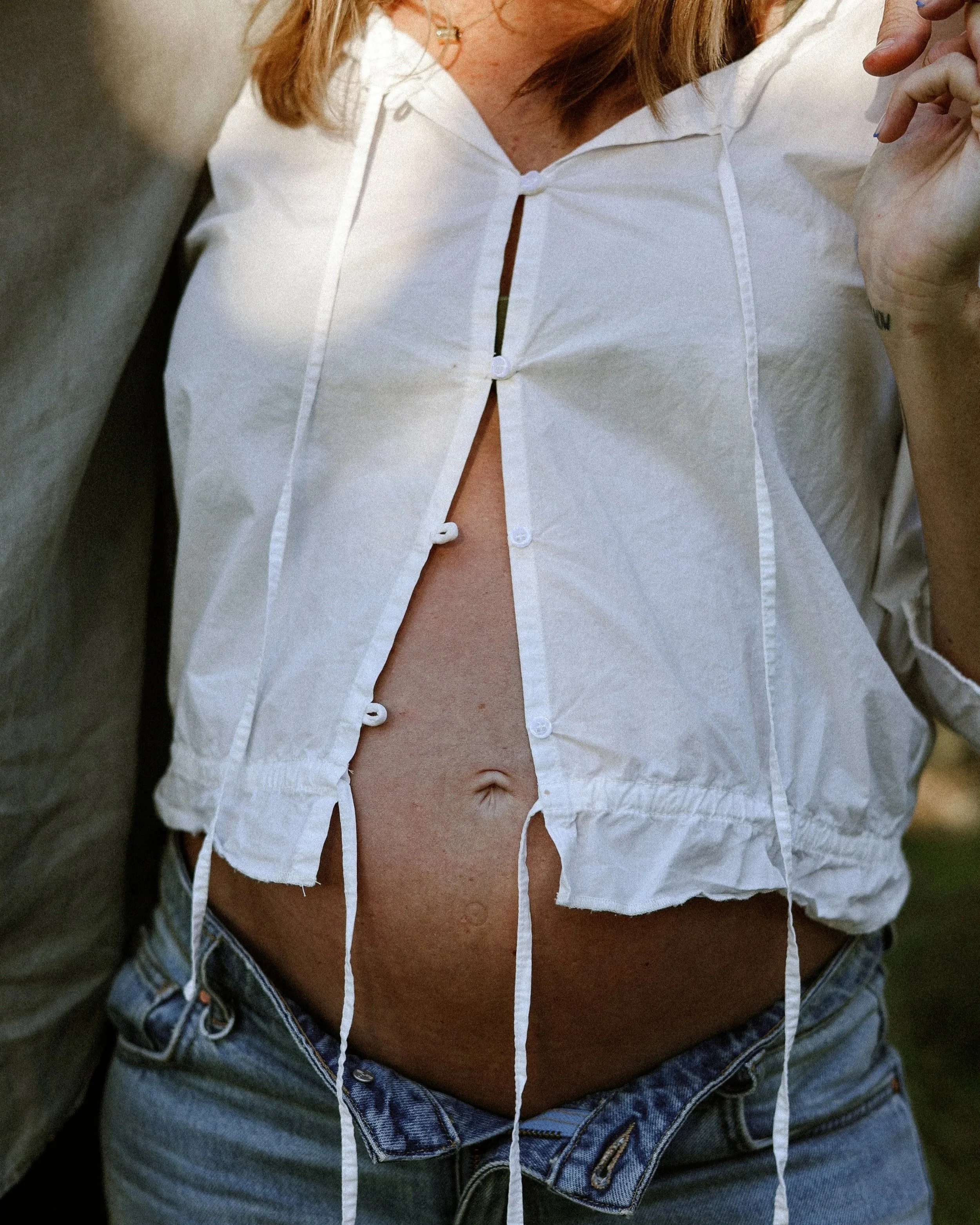A pregnant woman wearing a white shirt with buttons undone, revealing a portion of their chest, and blue denim jeans.