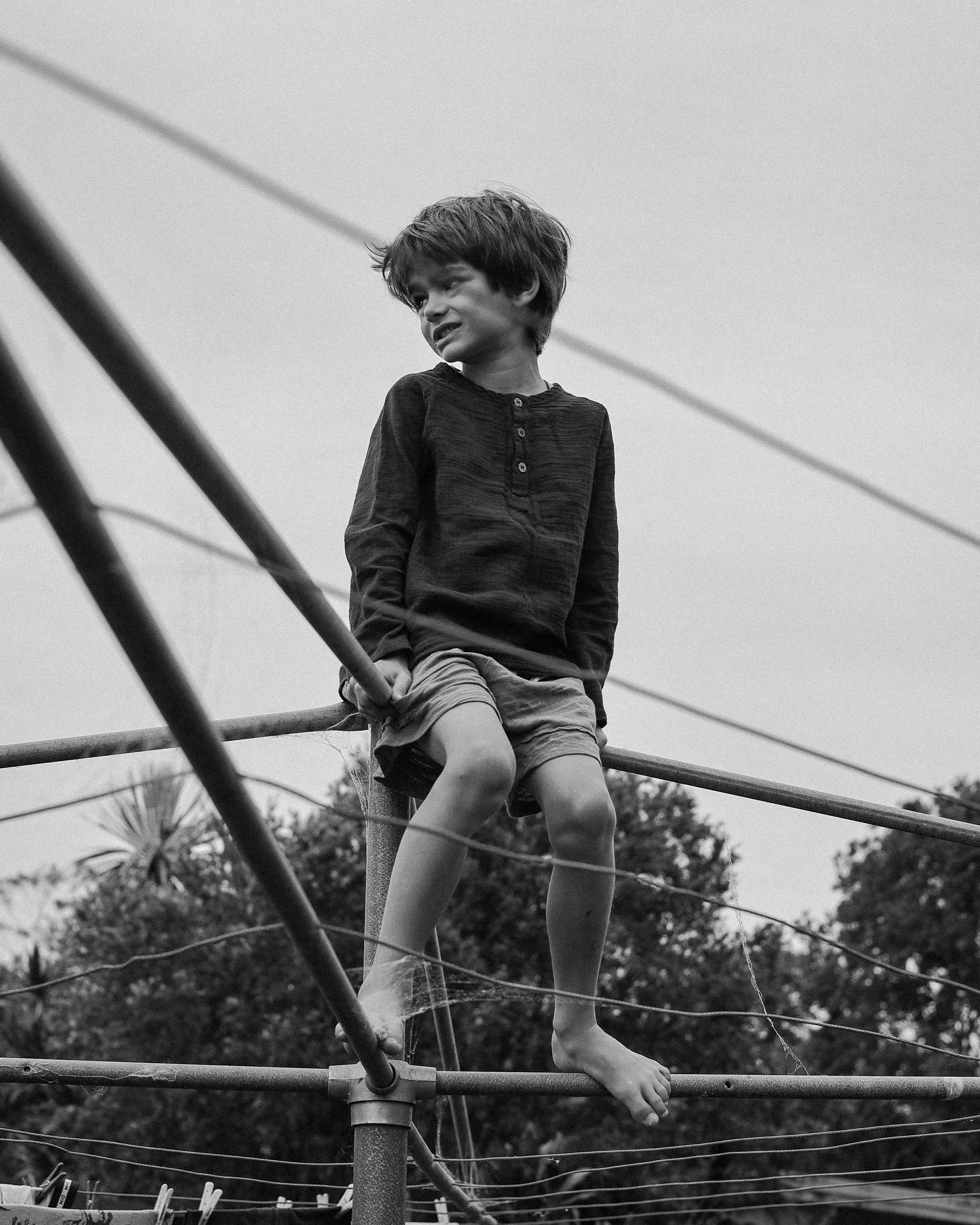 A black-and-white photograph of a young boy climbing on outdoor washing line. The boy is sitting on the structure with a clothbound top and is smiling while looking away. Trees are visible in the background.