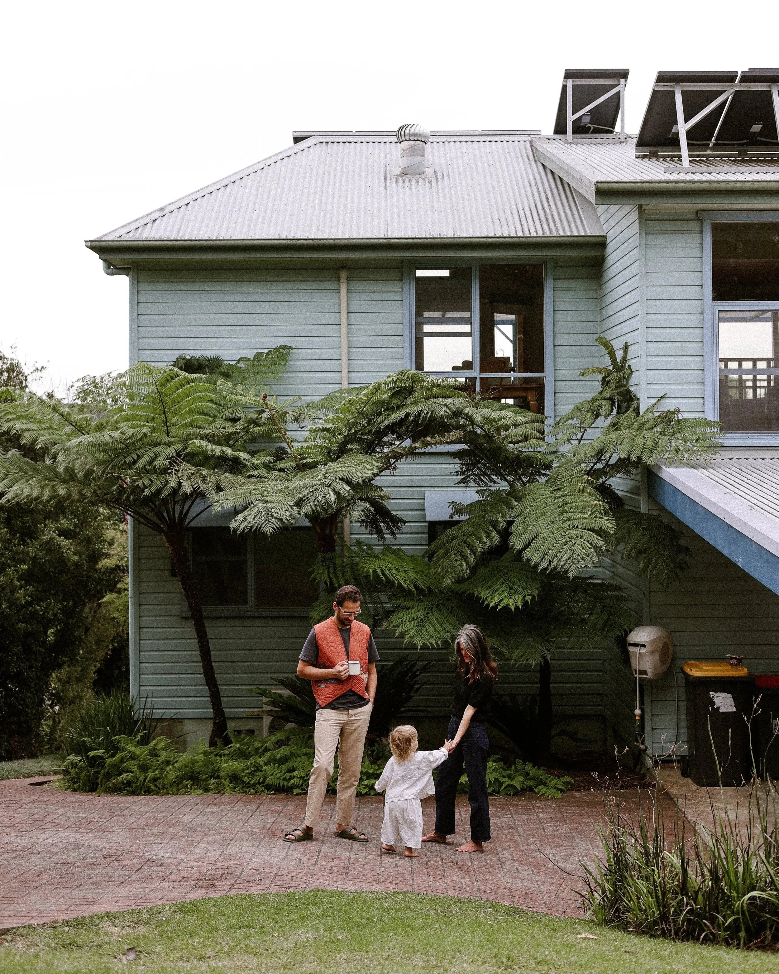 Family outside house, adults and a child talking on a brick patio, greenery around, house with solar panels on roof, large windows, some plants.