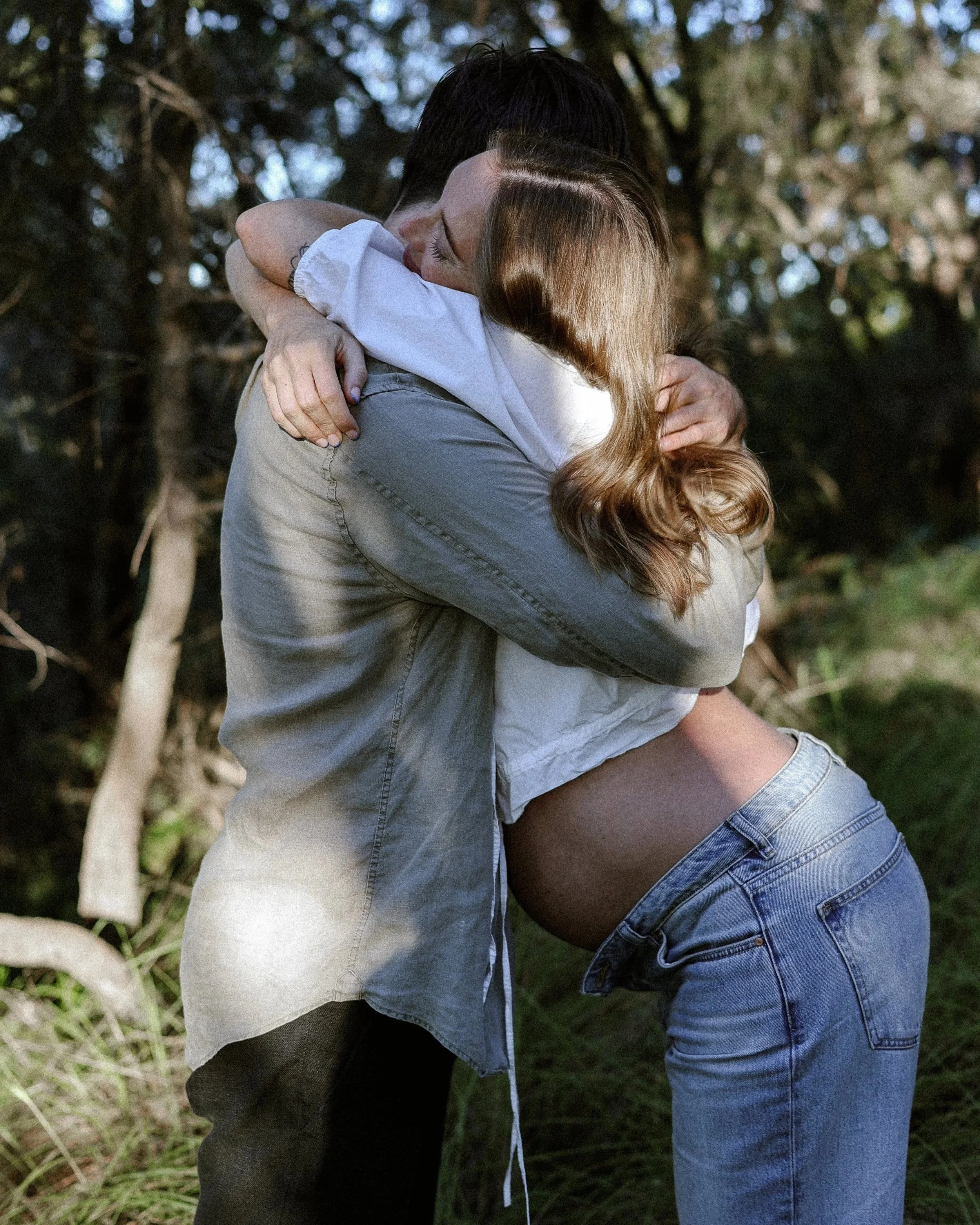 A man and his pregnant partner hugging outdoors in a forested area, with the woman facing away and the other man facing the camera, both with curly hair and casual clothing. The woman is 7 months pregnant.