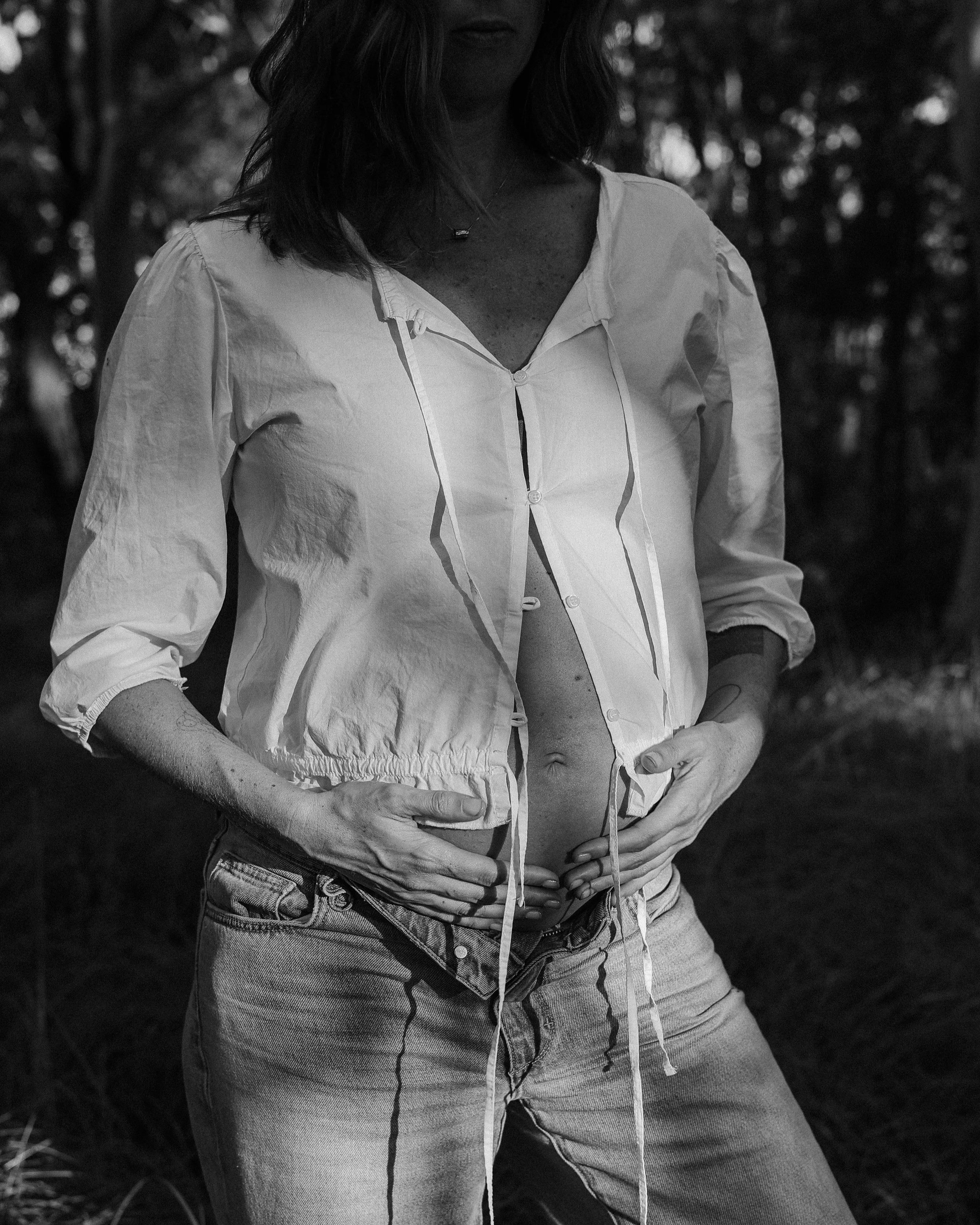 A pregnant mother-to-be wearing jeans and a white button-up shirt, standing with her hands on her hips outdoors, in a wooded area. The image is in black and white and the light is dappled.