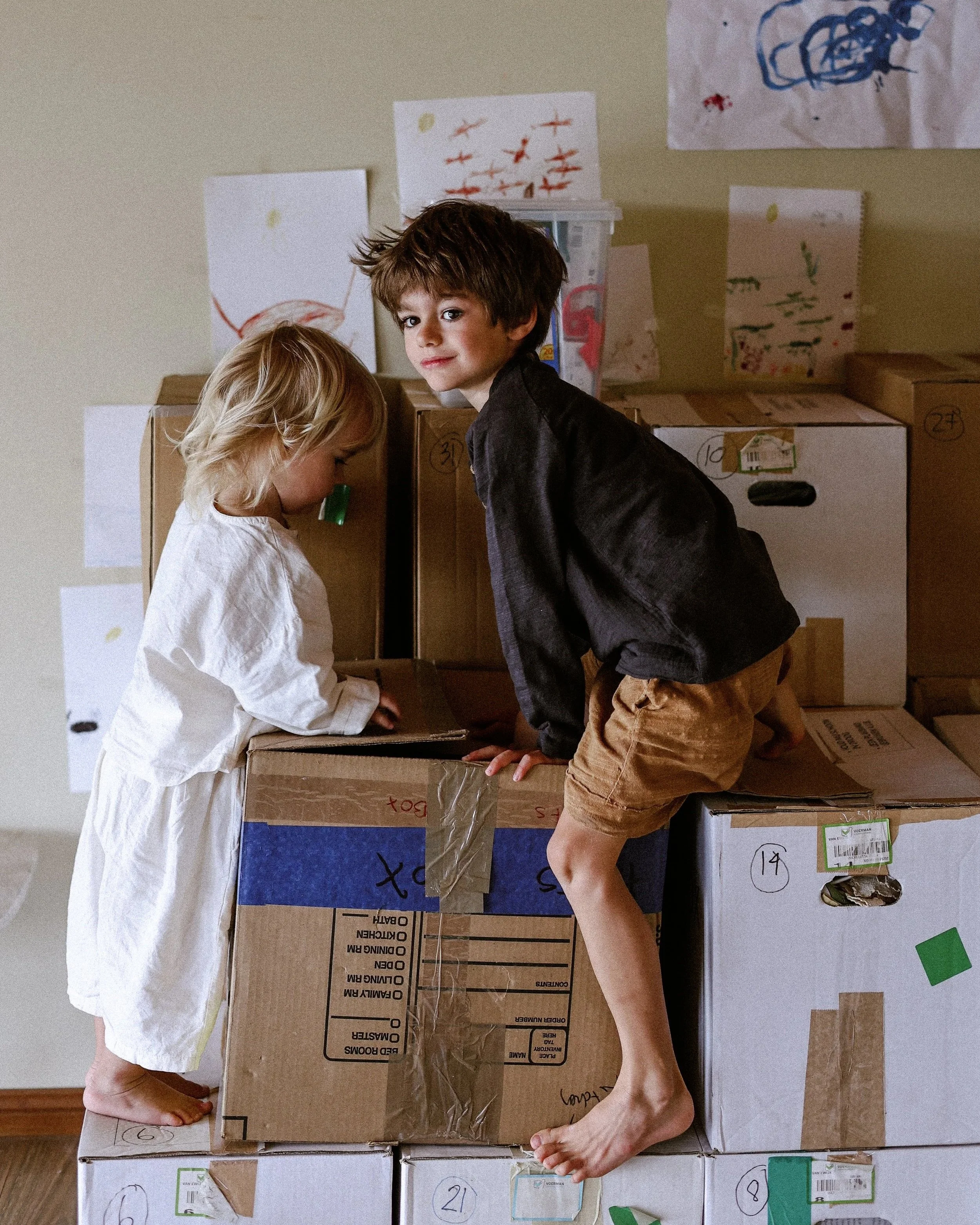 Two children, a girl and a boy, are kneeling on the floor among stacked cardboard boxes, with drawings and labels on them. The girl has blonde hair and is wearing a white dress, while the boy has brown hair, a black shirt, and brown shorts. The backg