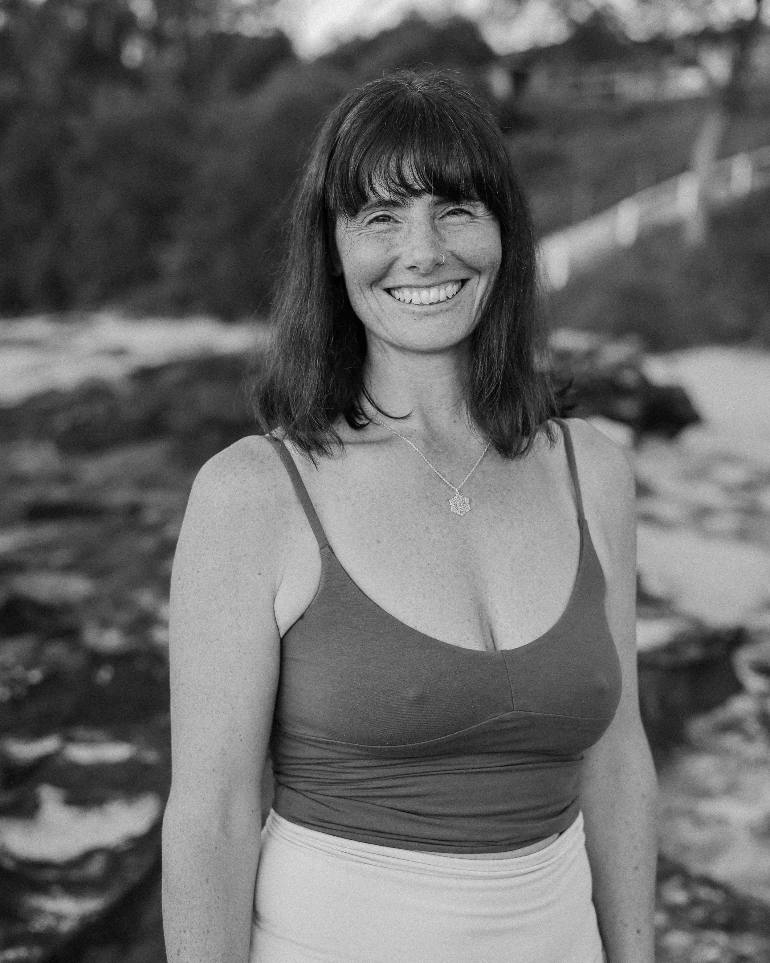 A woman with long dark hair, smiling, wearing a dark sleeveless top with thin straps, and a necklace with a decorative pendant.