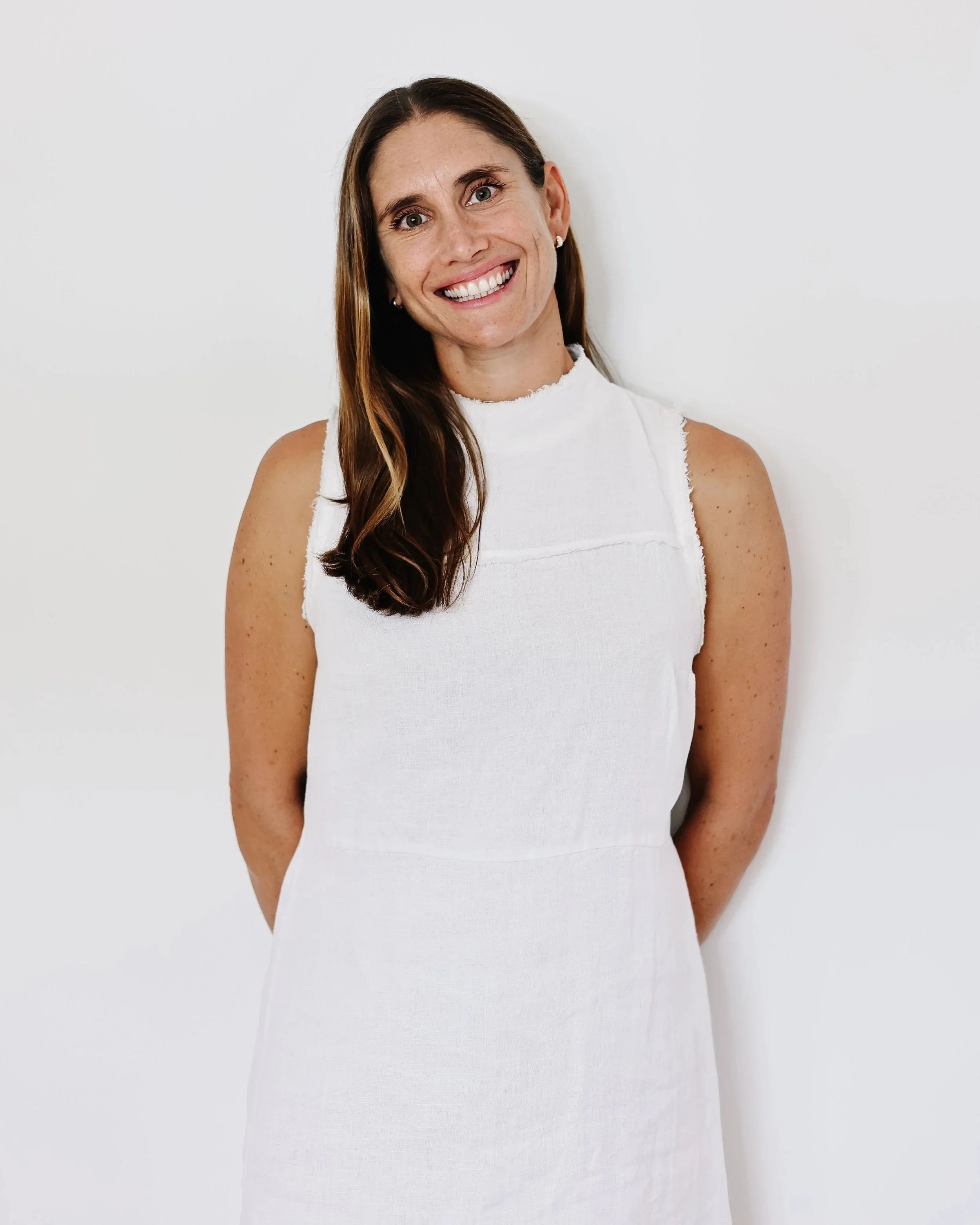 Portrait of a smiling woman with long brown hair, wearing a sleeveless white top, standing against a plain white background.