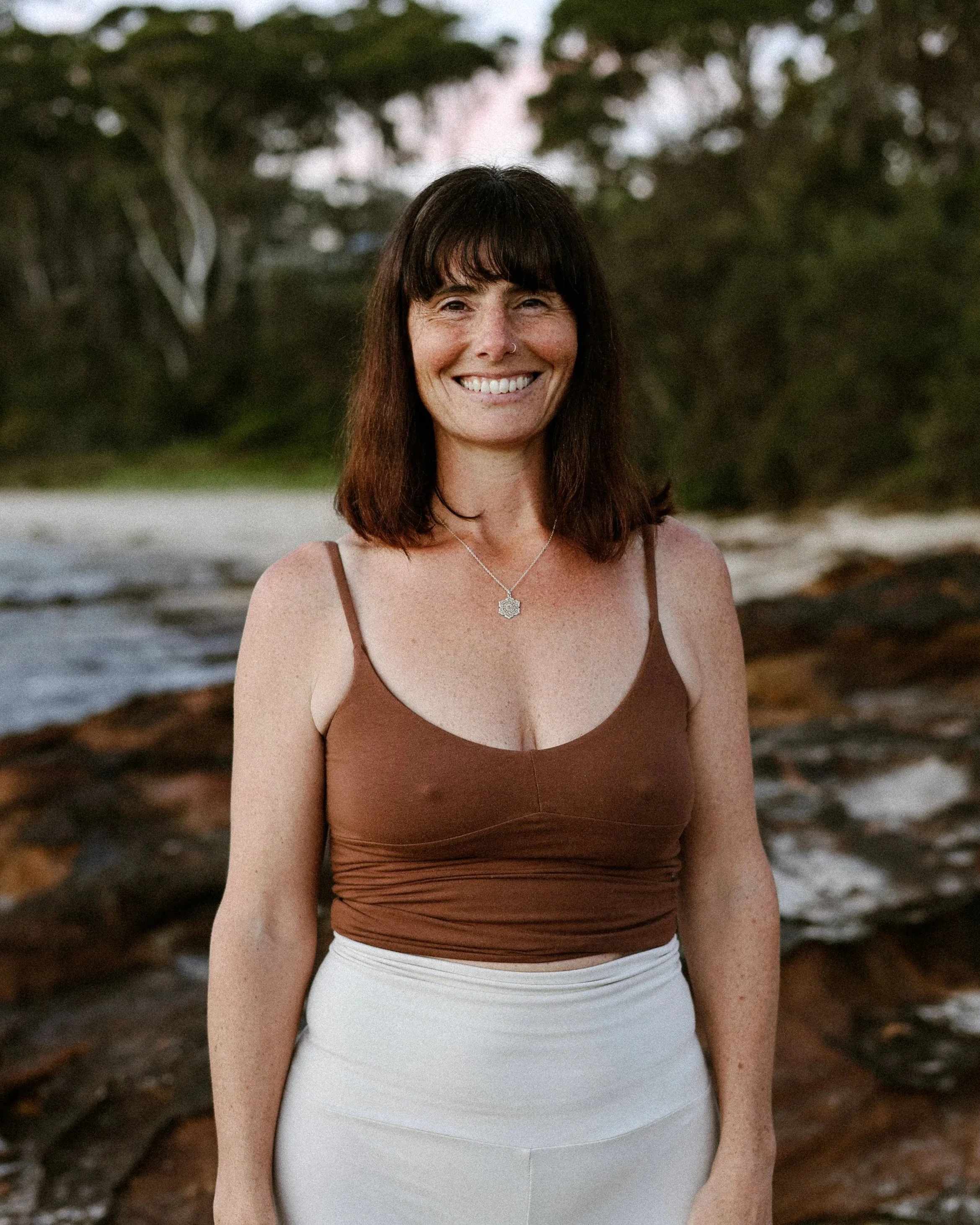 A woman with shoulder-length dark brown hair, light skin, and freckles, smiling at the camera, standing outdoors near a beach with trees and water in the background.