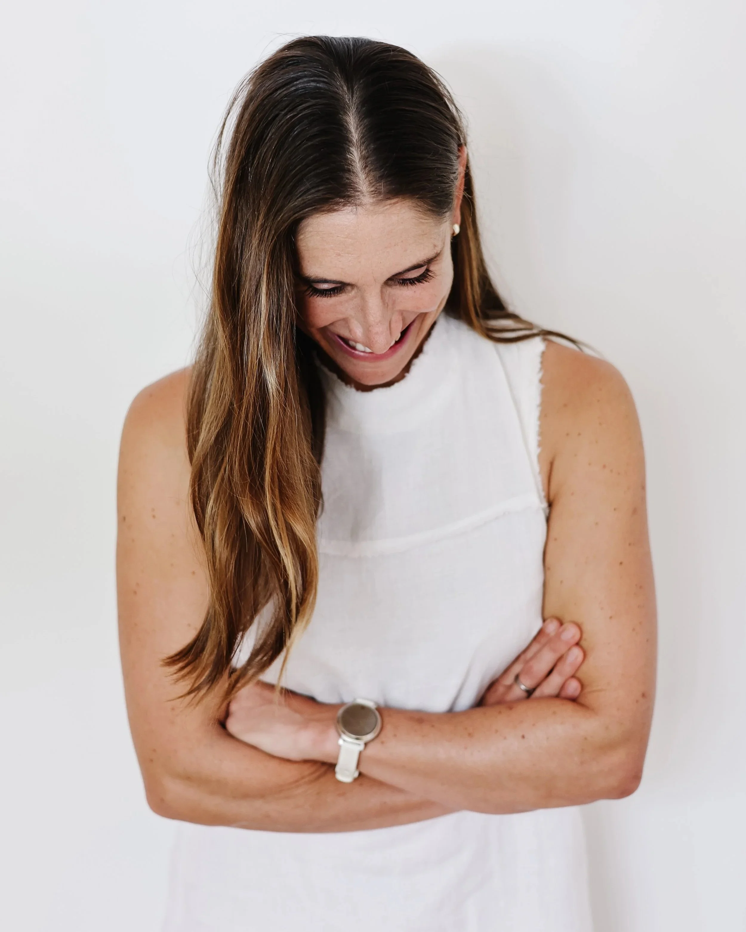 A woman with long brown hair sharing a smile while looking down wearing a white sleeveless top and a watch on her wrist, standing against a plain white background.